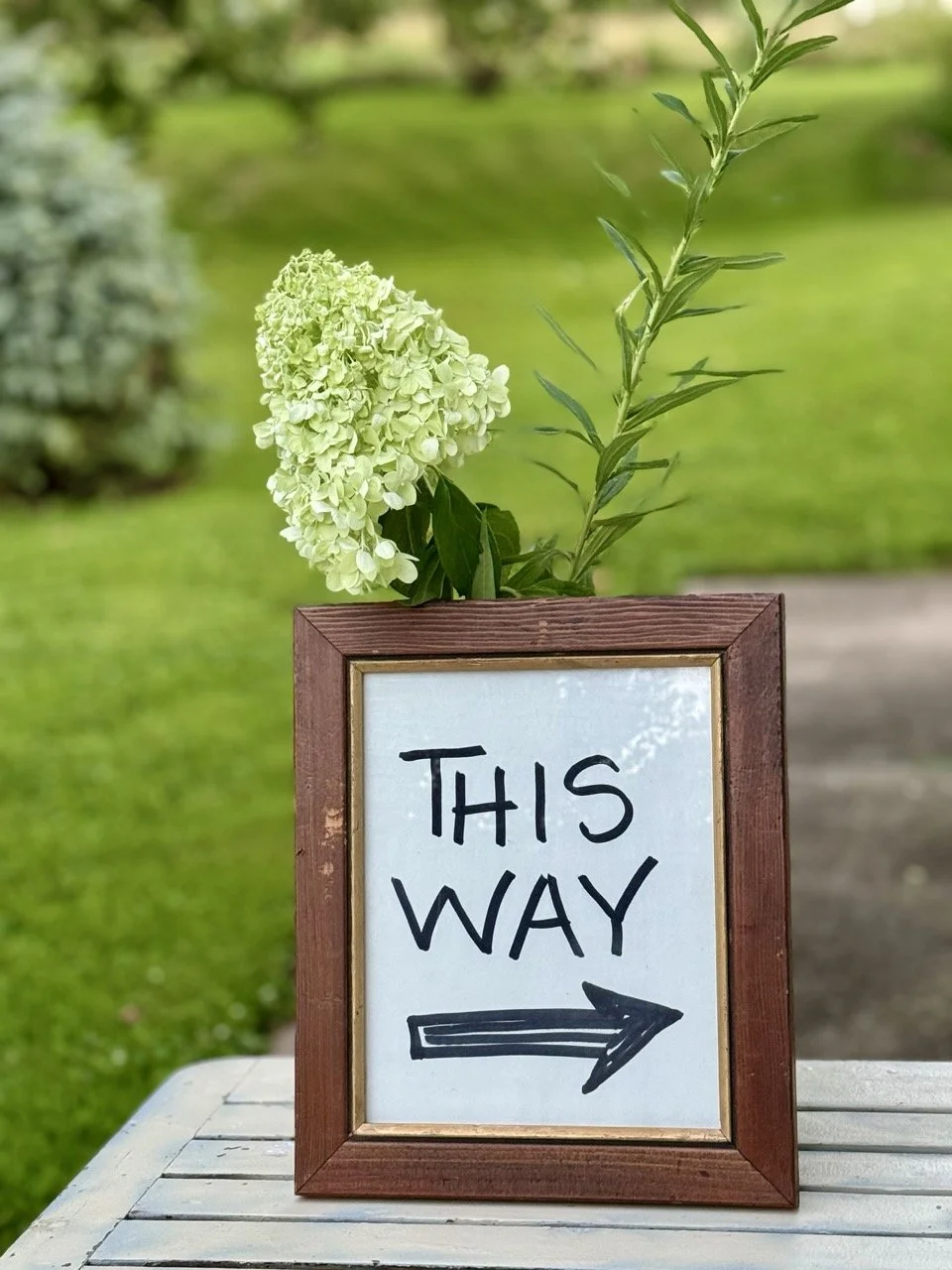 A wooden sign with a white background and black handwritten text saying "This Way" and an arrow pointing to the right. The sign is decorated with a small bouquet of green and white flowers attached to the top, placed on a white wooden surface outdoor