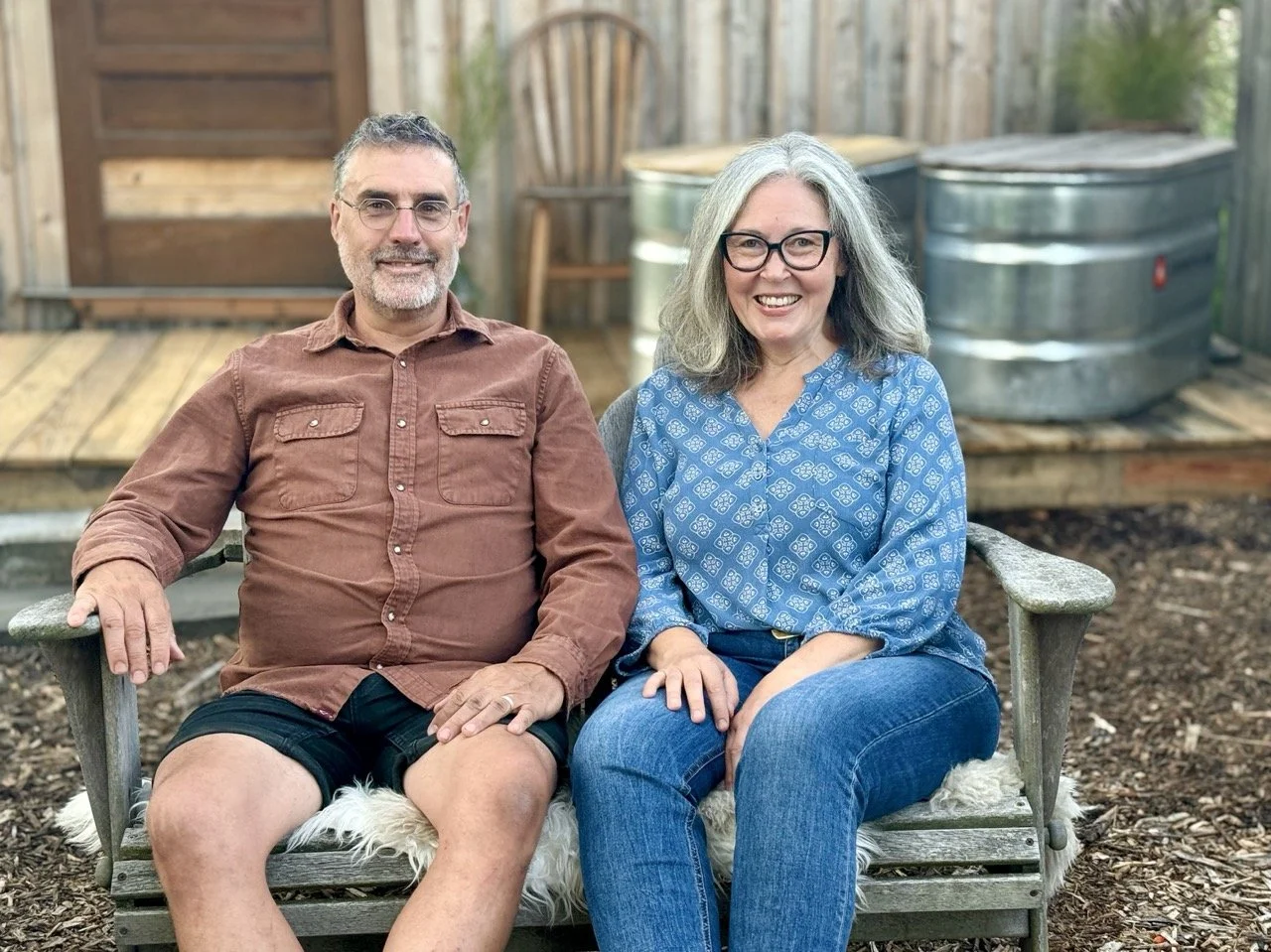 A smiling man and woman sitting on a wooden bench outdoors, with a wooden deck, a wooden fence, and two metal water tanks in the background.
