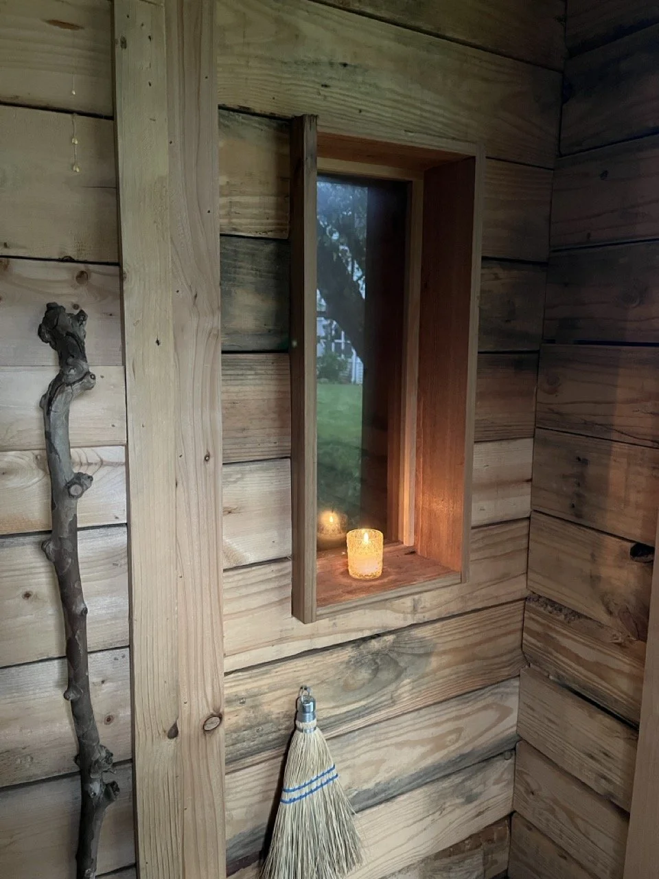 Wood-paneled wall with a small rectangular window and flickering candle on the windowsill, a small branch as the handle of a wooden door, and a broom hanging from a nail on the wall.
