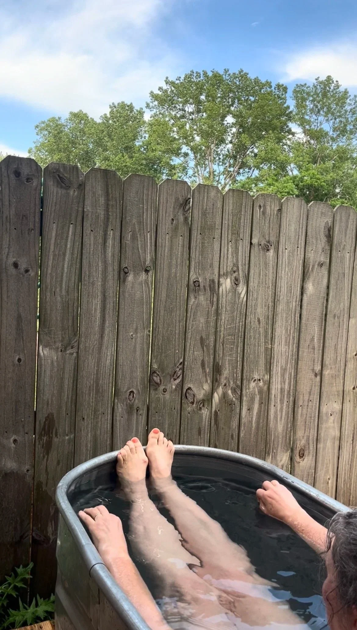 Person relaxing in an outdoor hot tub with legs and feet visible, pool water, and a wooden privacy fence. Background includes green trees and a blue sky with some clouds.