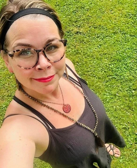 A woman with glasses and a black headband taking a selfie outdoors on green grass, wearing a black tank top and layered necklaces.