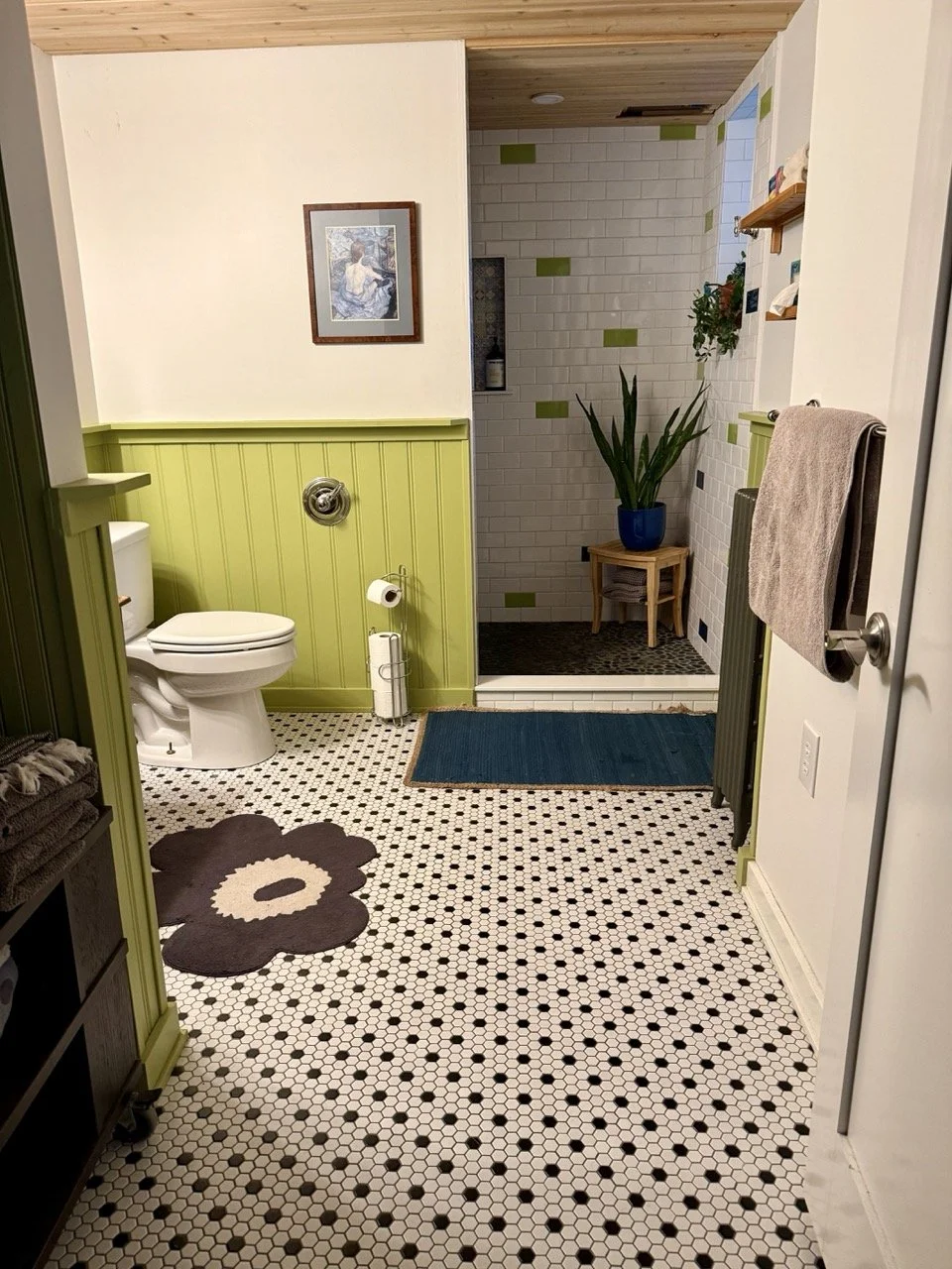 Bathroom with green and white paneled walls, black and white hexagon tile floor, a toilet, a blue rug, a large potted plant, and a walk-in shower with white brick tiles and green accents.