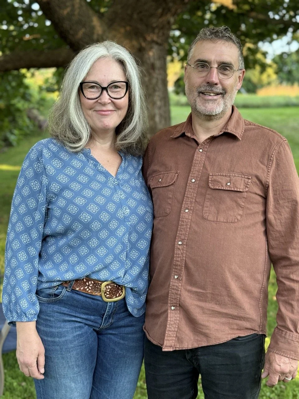 A middle-aged woman with gray hair and glasses wearing a blue patterned blouse and jeans, standing next to a middle-aged man with glasses and a beard, wearing a brown button-up shirt and black pants, in a park with trees in the background.