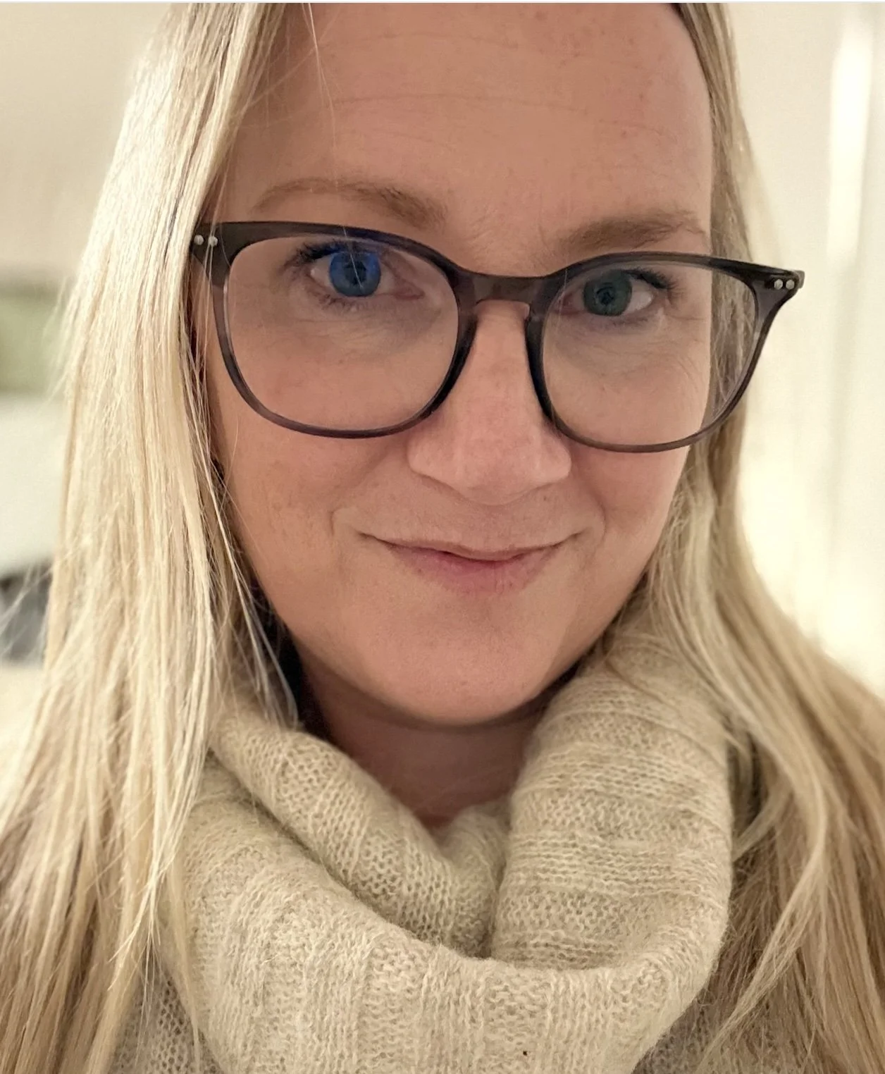 A close-up photo of a woman with blonde hair wearing glasses and a beige knitted scarf, smiling gently.