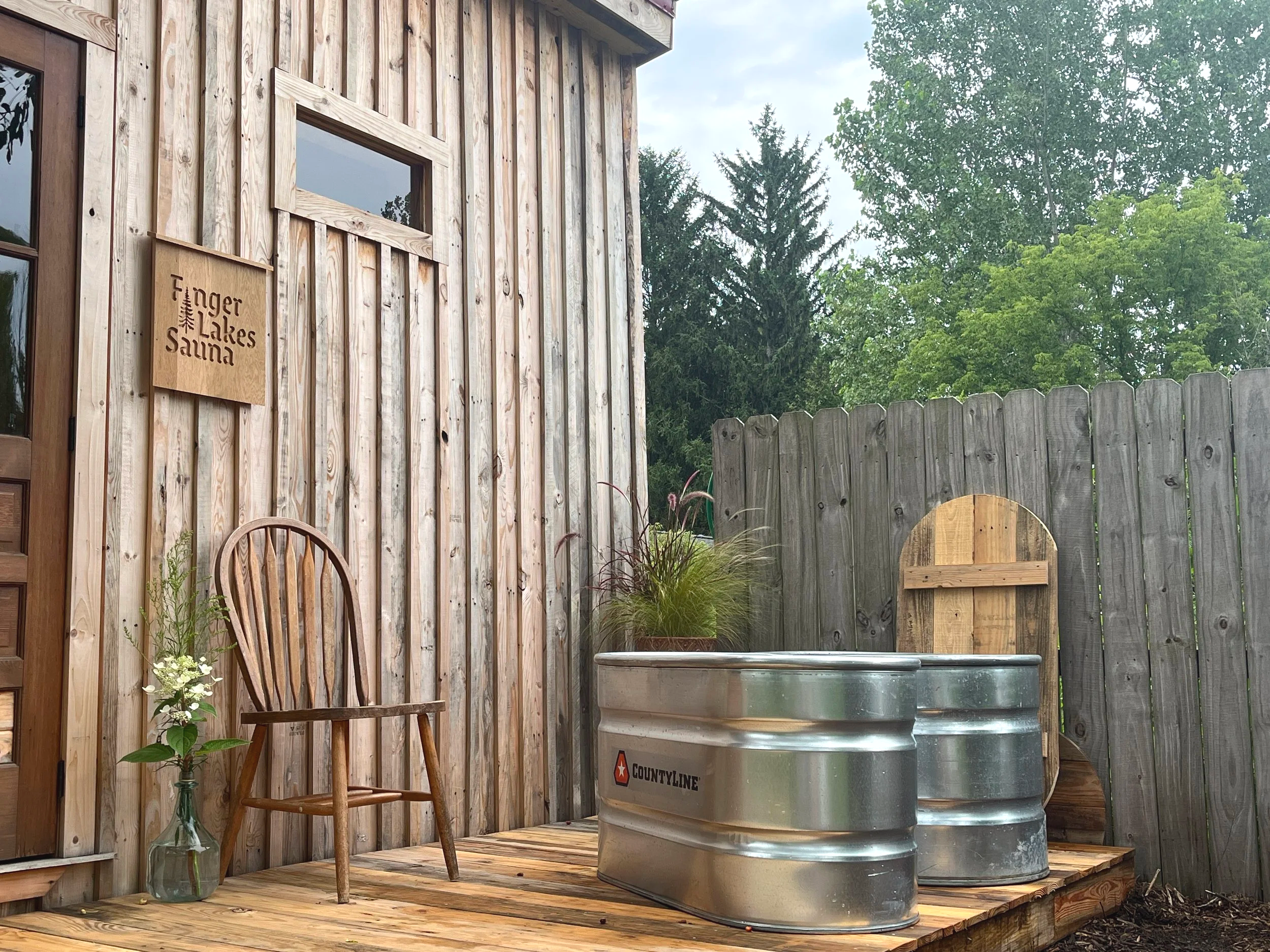 A wooden deck with a wooden chair, a glass vase with flowers, and two metal tubs on a wooden platform. A wooden fence and trees are in the background. There is a sign that reads "Finger Lakes Sauna" on the wooden exterior of a sauna.