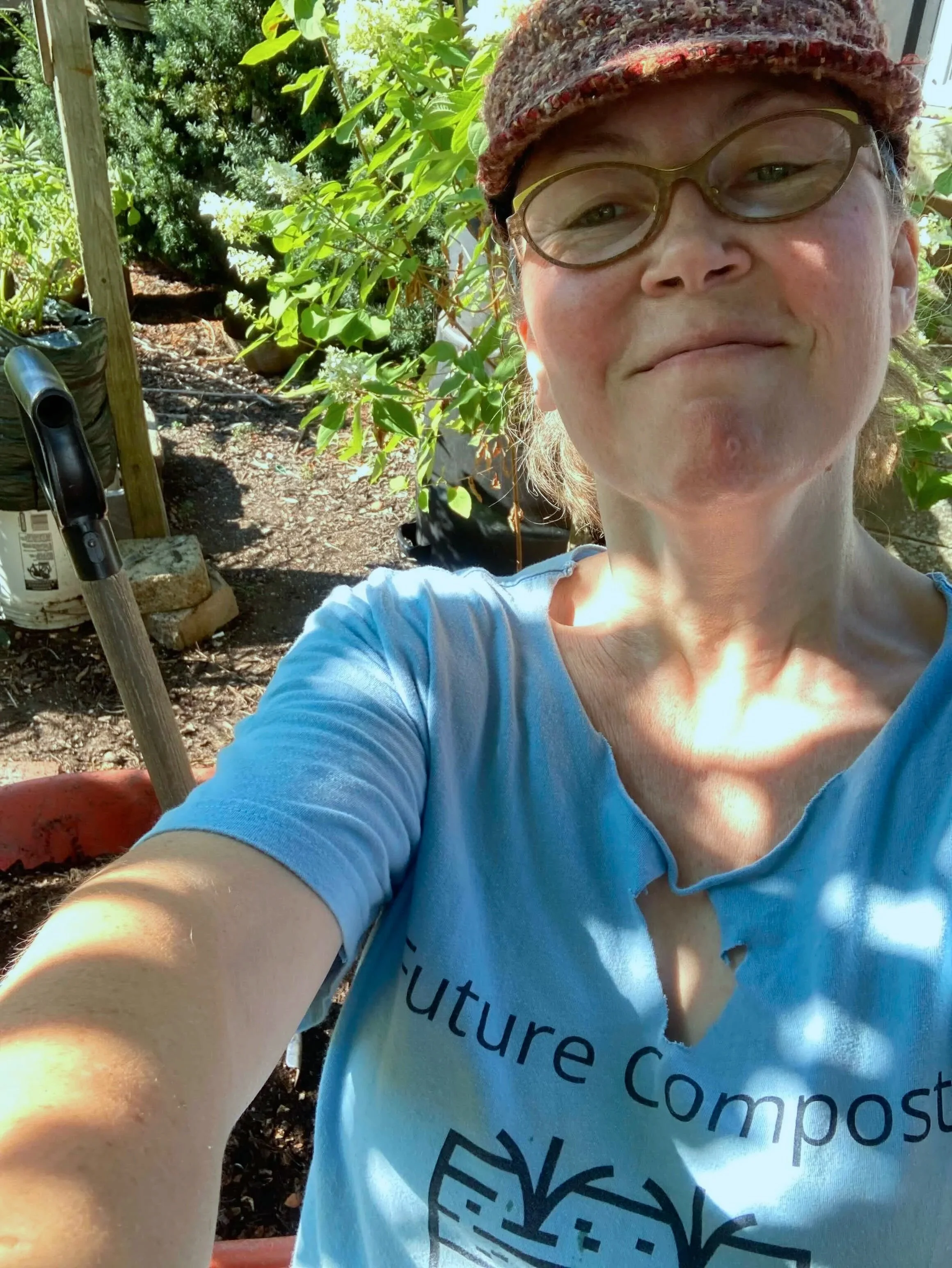 A woman with glasses and a multicolored hat taking a selfie outdoors, with greenery, plants, and gardening tools visible in the background.