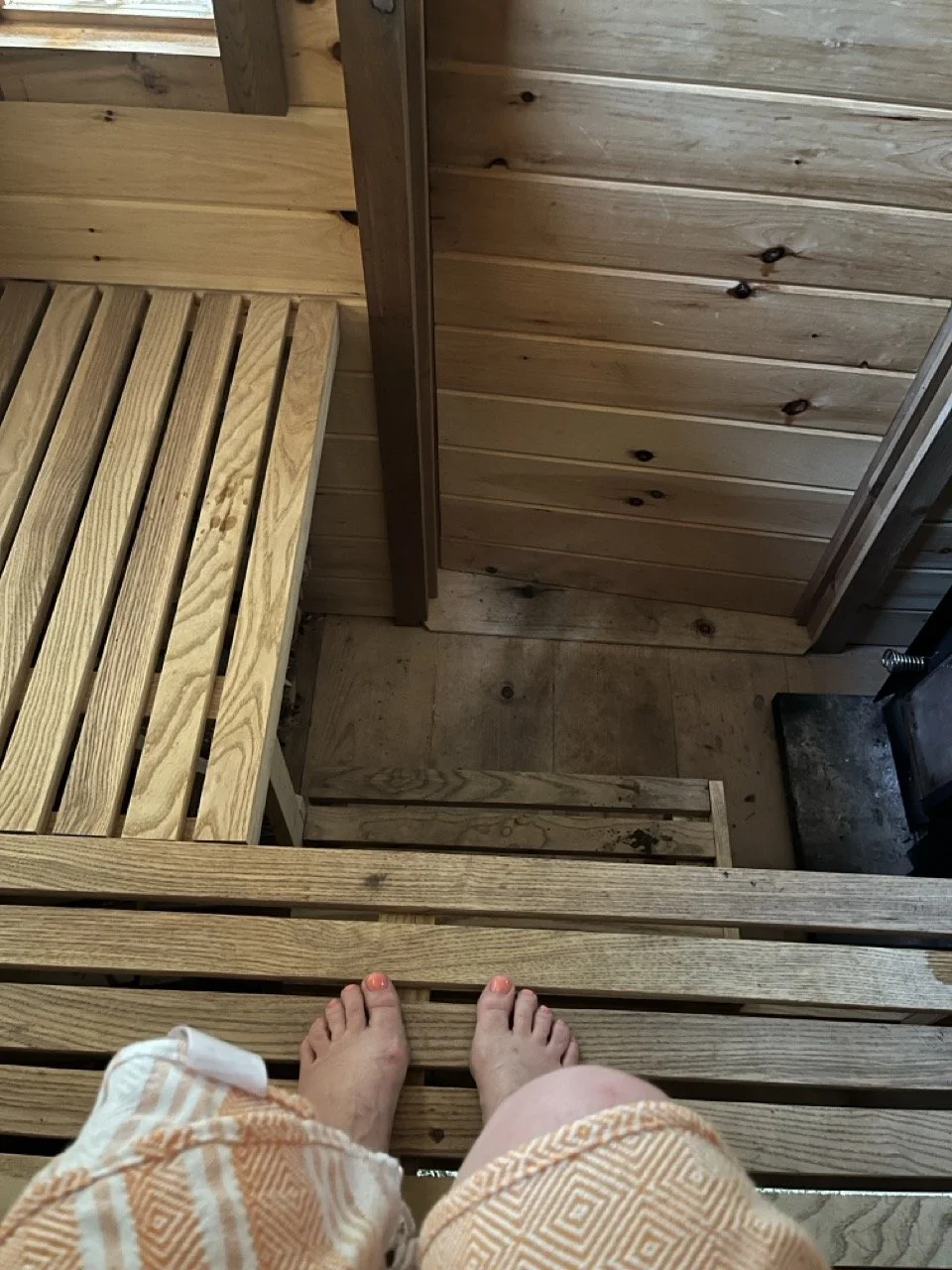 View from a wooden bench inside a sauna, showing a person's legs and feet with toenails painted orange as they sit on the bench, looking down at the wooden benches, floor and door.