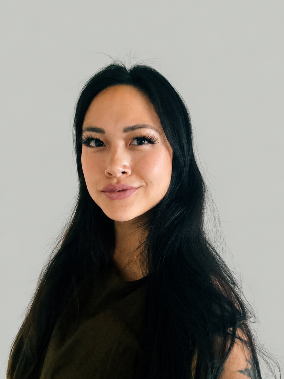 Portrait of a woman with long black hair, wearing a dark top, standing against a plain background.