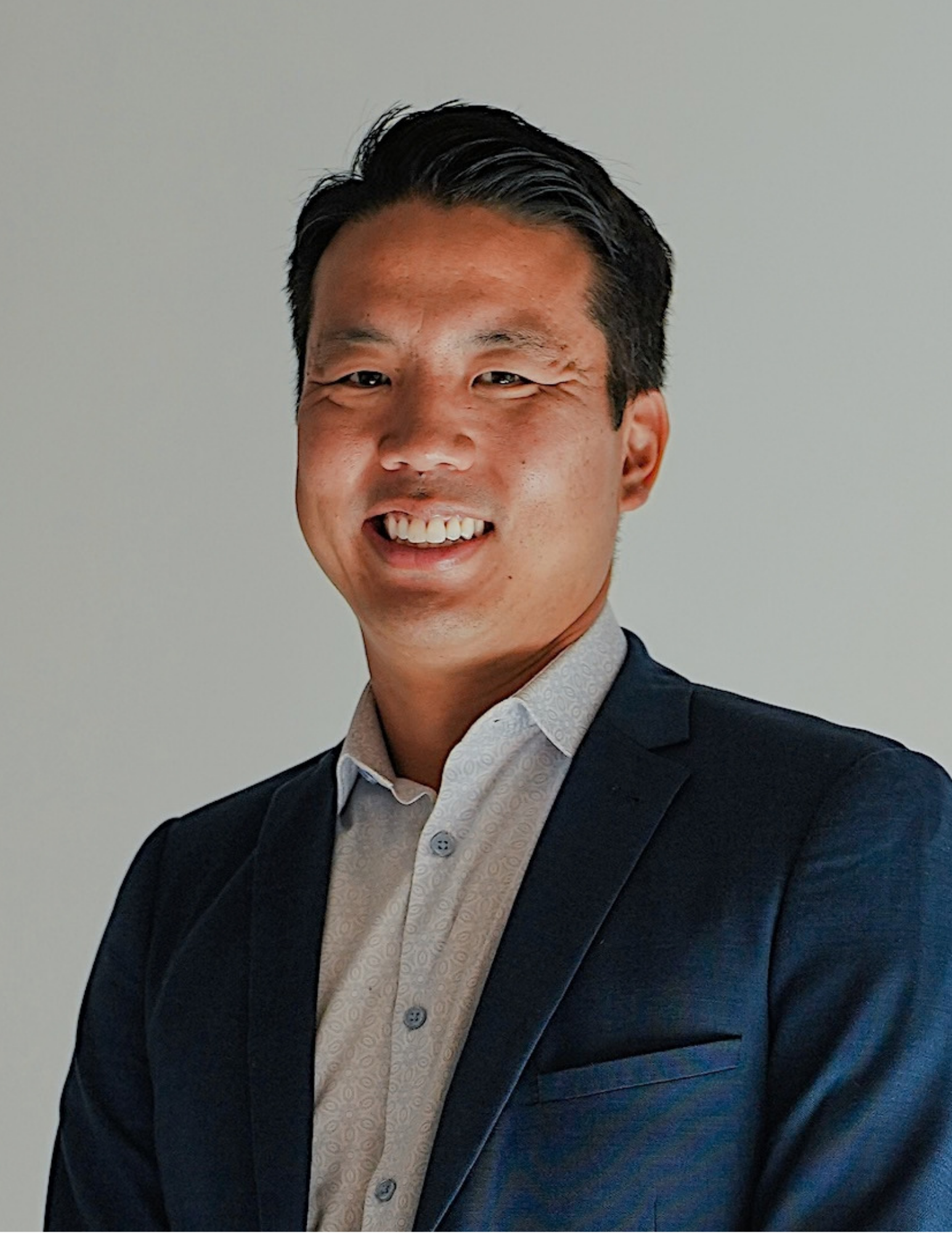 A smiling man in a business suit with a white textured shirt standing against a plain light wall.