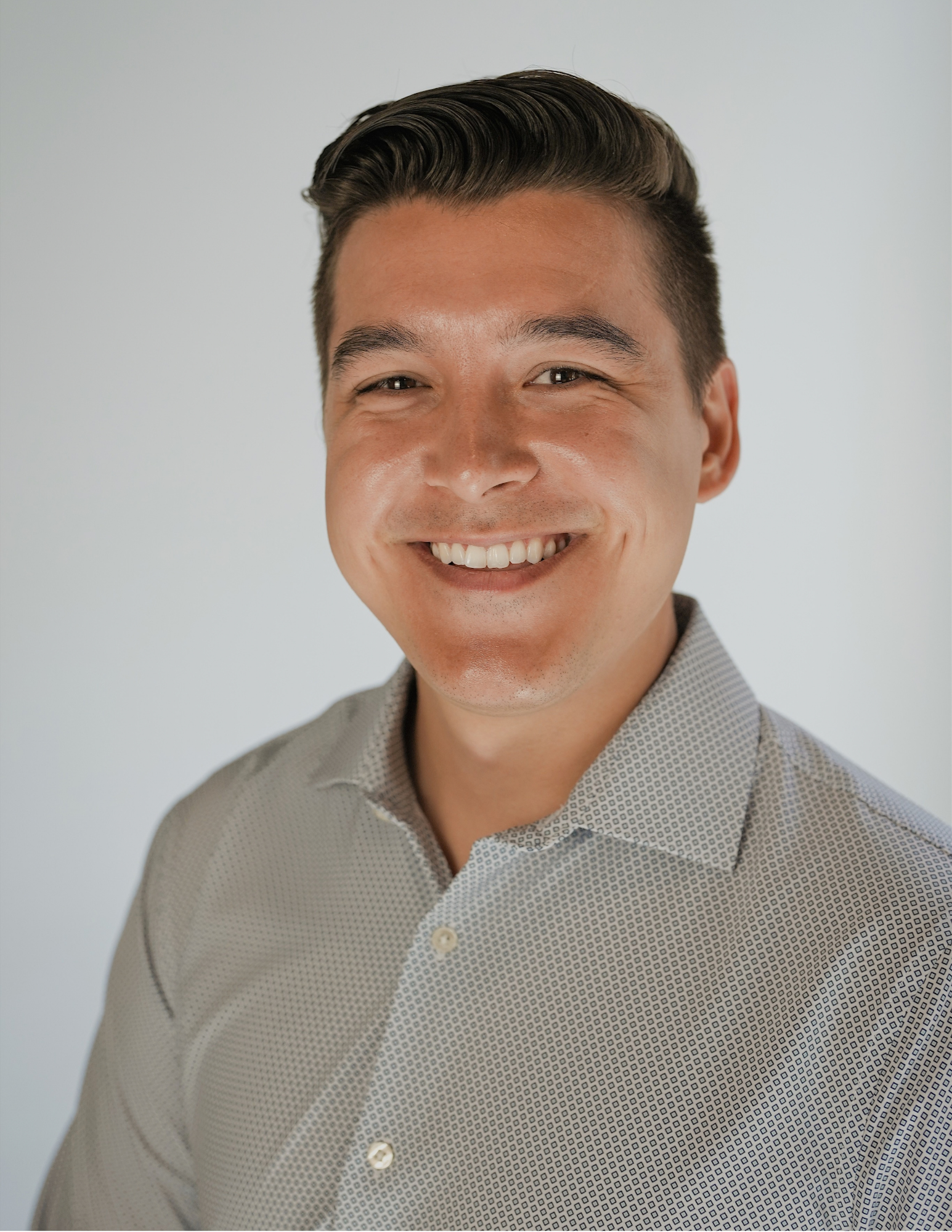 Headshot of a young man with dark hair styled to the side, smiling, wearing a light gray patterned button-up shirt, against a plain light background.