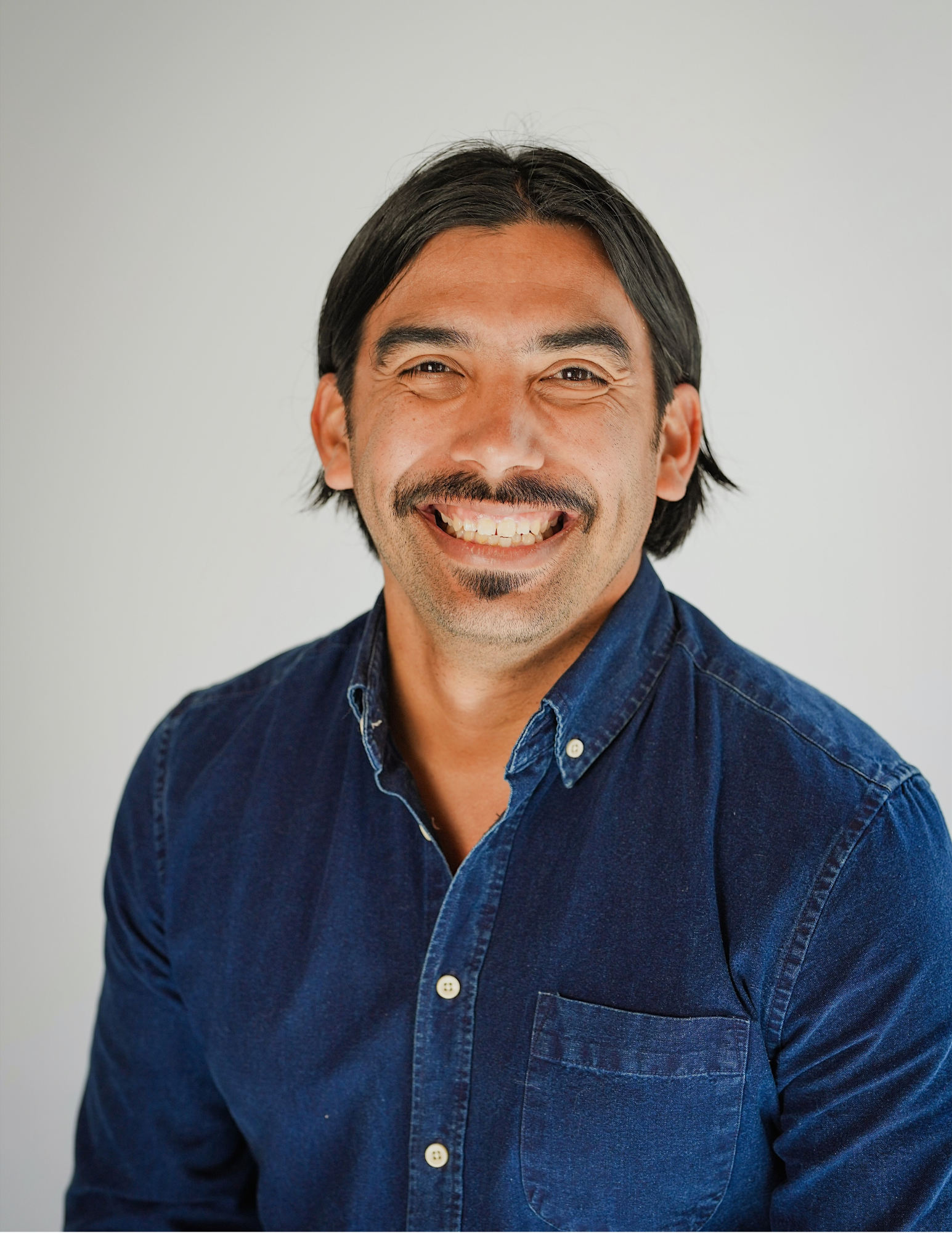 Close-up portrait of a smiling man with dark hair and a mustache wearing a dark blue denim shirt against a plain light gray background.