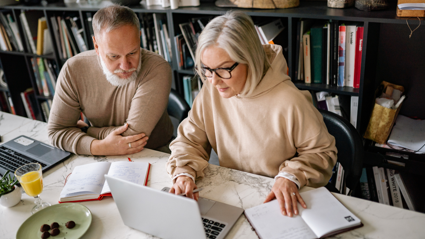 A man and woman sitting at a table with a laptop, open notebooks, a glass of orange juice, and a plate of chocolate truffles, working together in an office with bookshelves in the background.