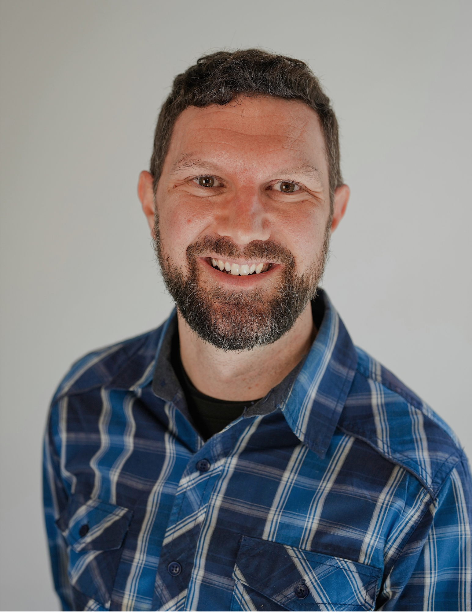 A smiling man with a beard and mustache wearing a blue plaid shirt, looking directly at the camera, standing against a plain light background. Jeremy Siegel, licensed insurance agent with Benefits NW.