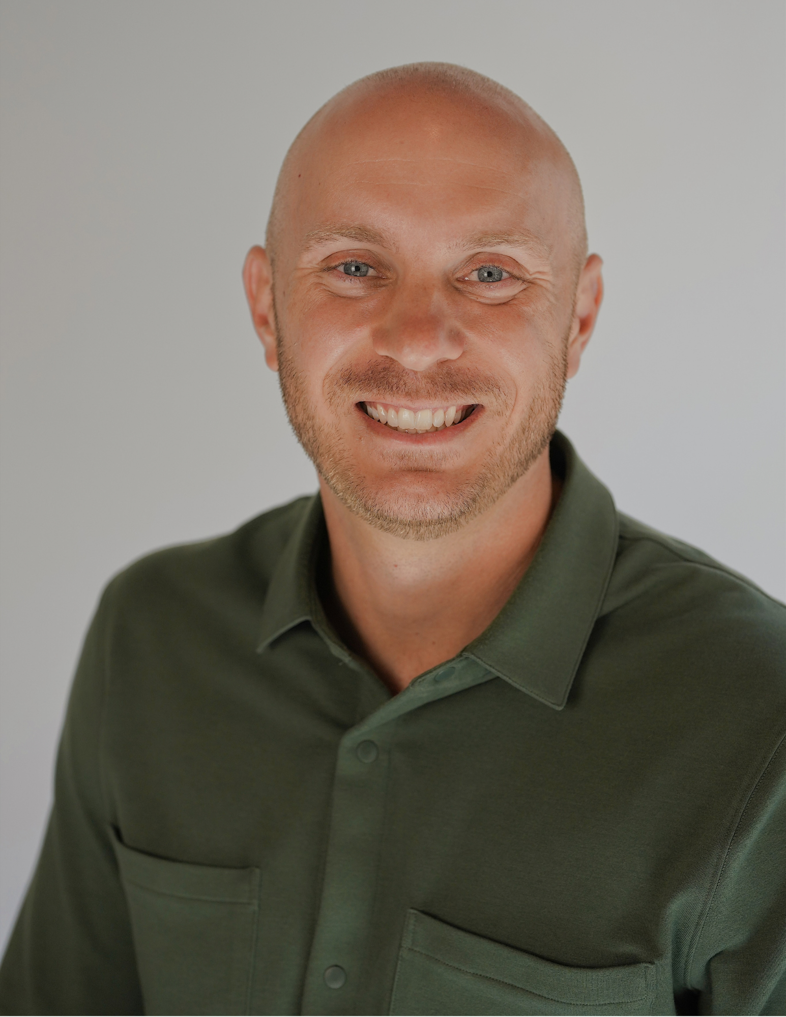 A close-up of a smiling man with a bald head, light skin, and short facial hair, wearing a dark green collared shirt against a plain light gray background. Michael Fish, licensed insurance agent with Benefits NW.