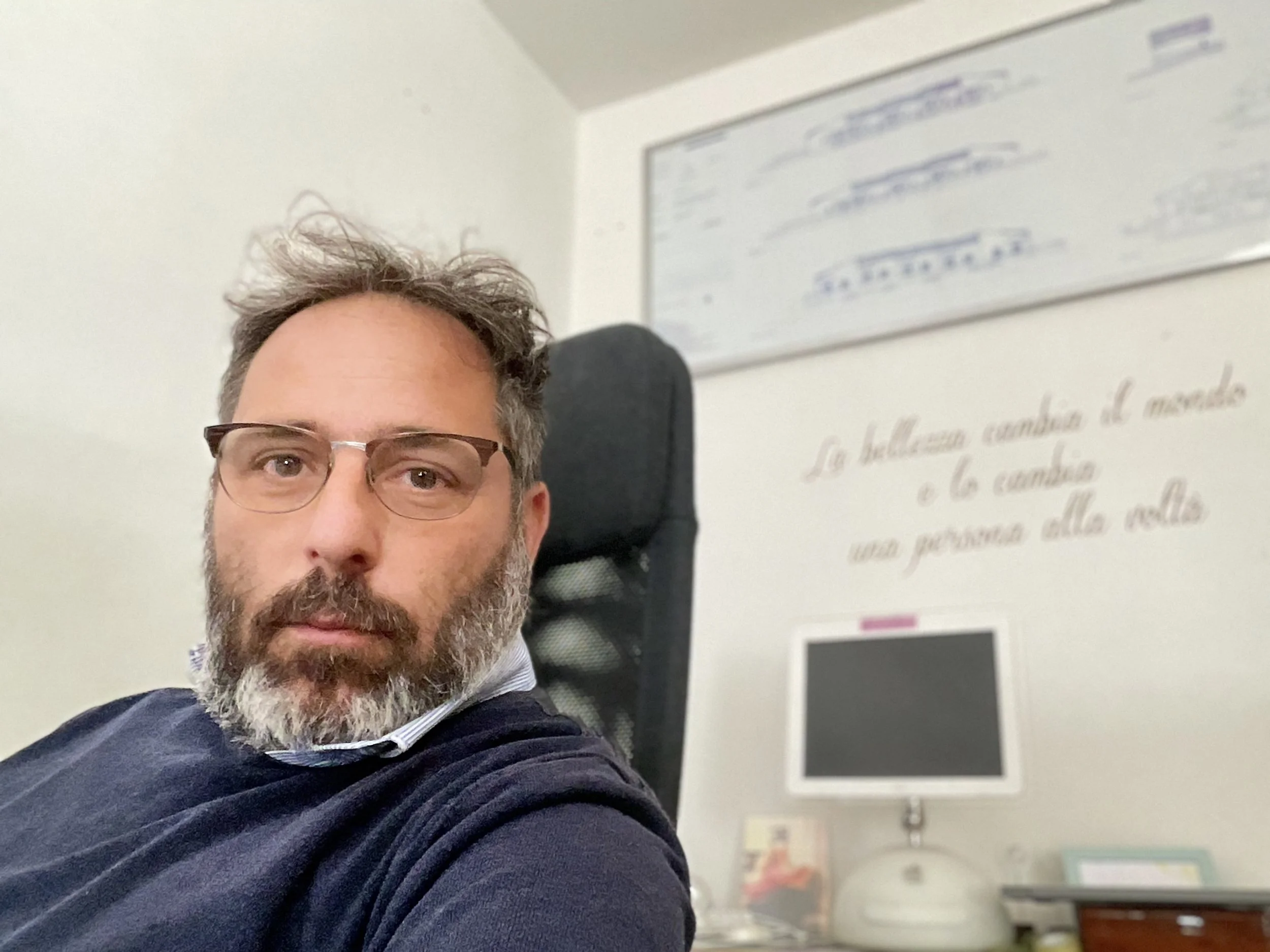 Un uomo con barba e capelli castani, indossa occhiali e una felpa blu, seduto su una sedia di lavoro in un ufficio. Sullo sfondo si vede una parete bianca con alcune cornici e scritte.