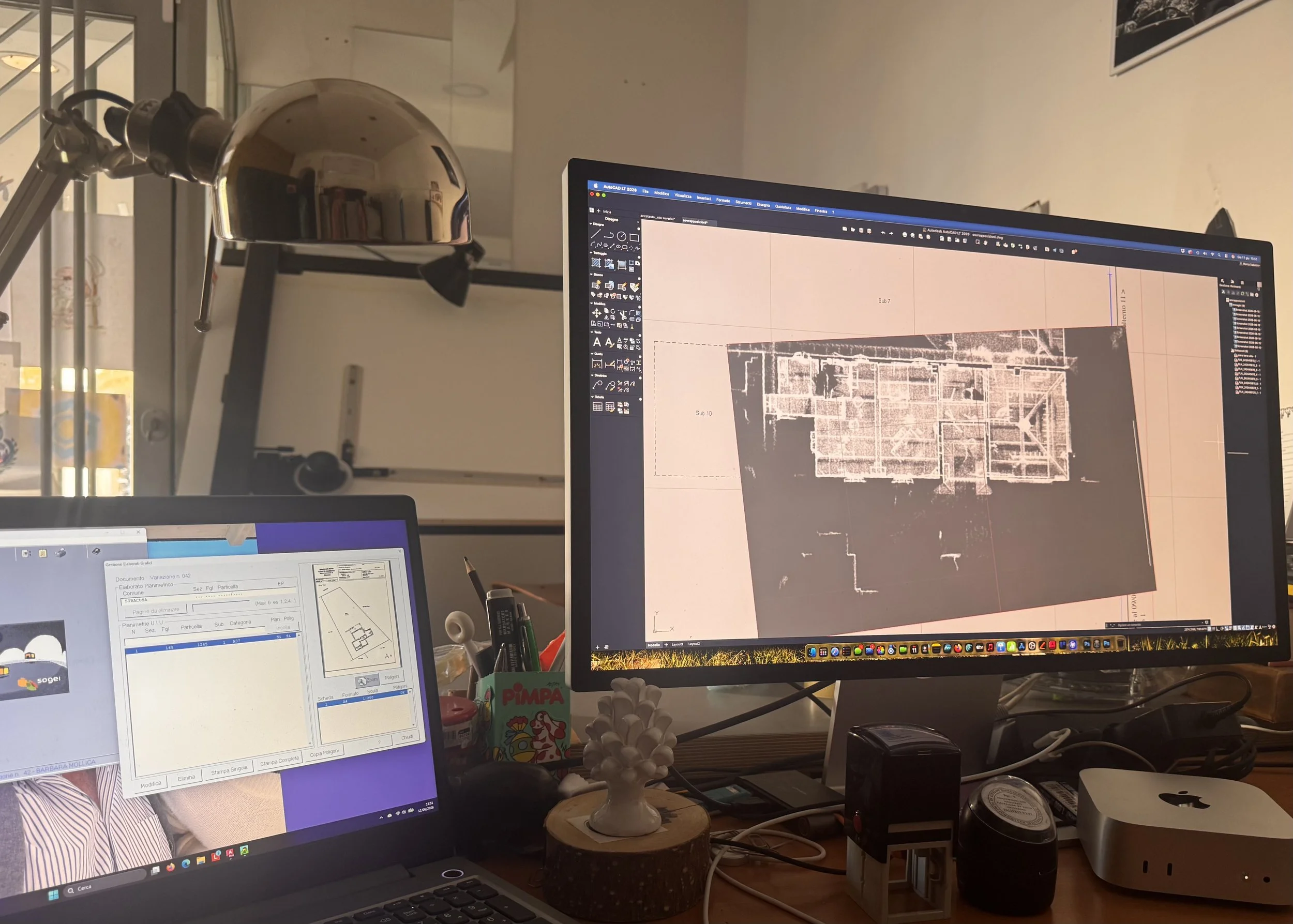 Postazione di lavoro con due monitor. Sul monitor principale si visualizza un software di progettazione con un piano architettonico, mentre sul secondo monitor si vede un documento di testo. La scrivania ha vari oggetti tra cui una pianta decorativa, penne, una scatola di PIMPA e altri elementi di cancelleria.