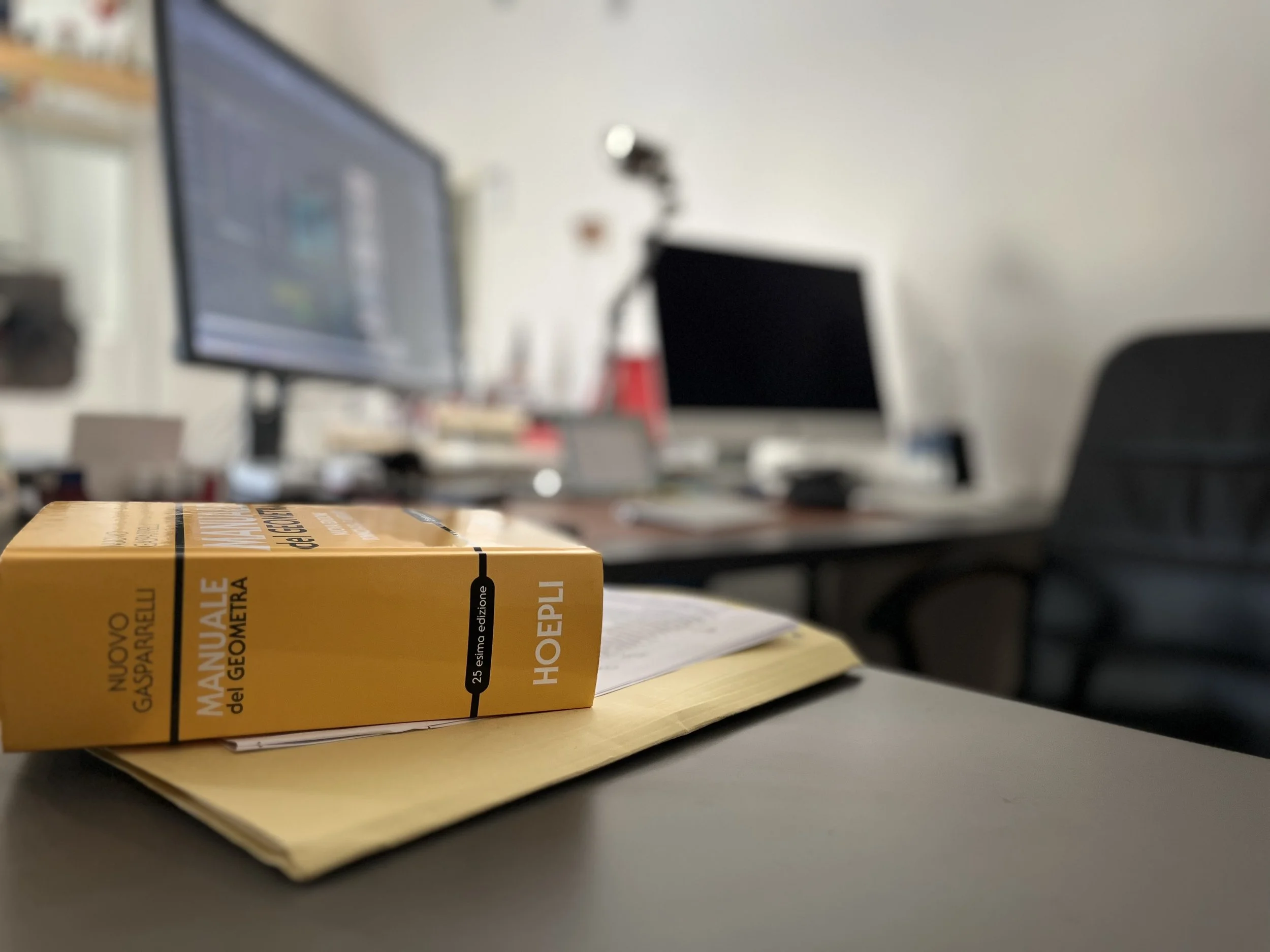 Prima piano di un tavolo con un libro giallo intitolato "Manuale da Geometra" e una cartellina sotto, in sfondo una scrivania con computer e altri oggetti, scena di un ufficio o studio.