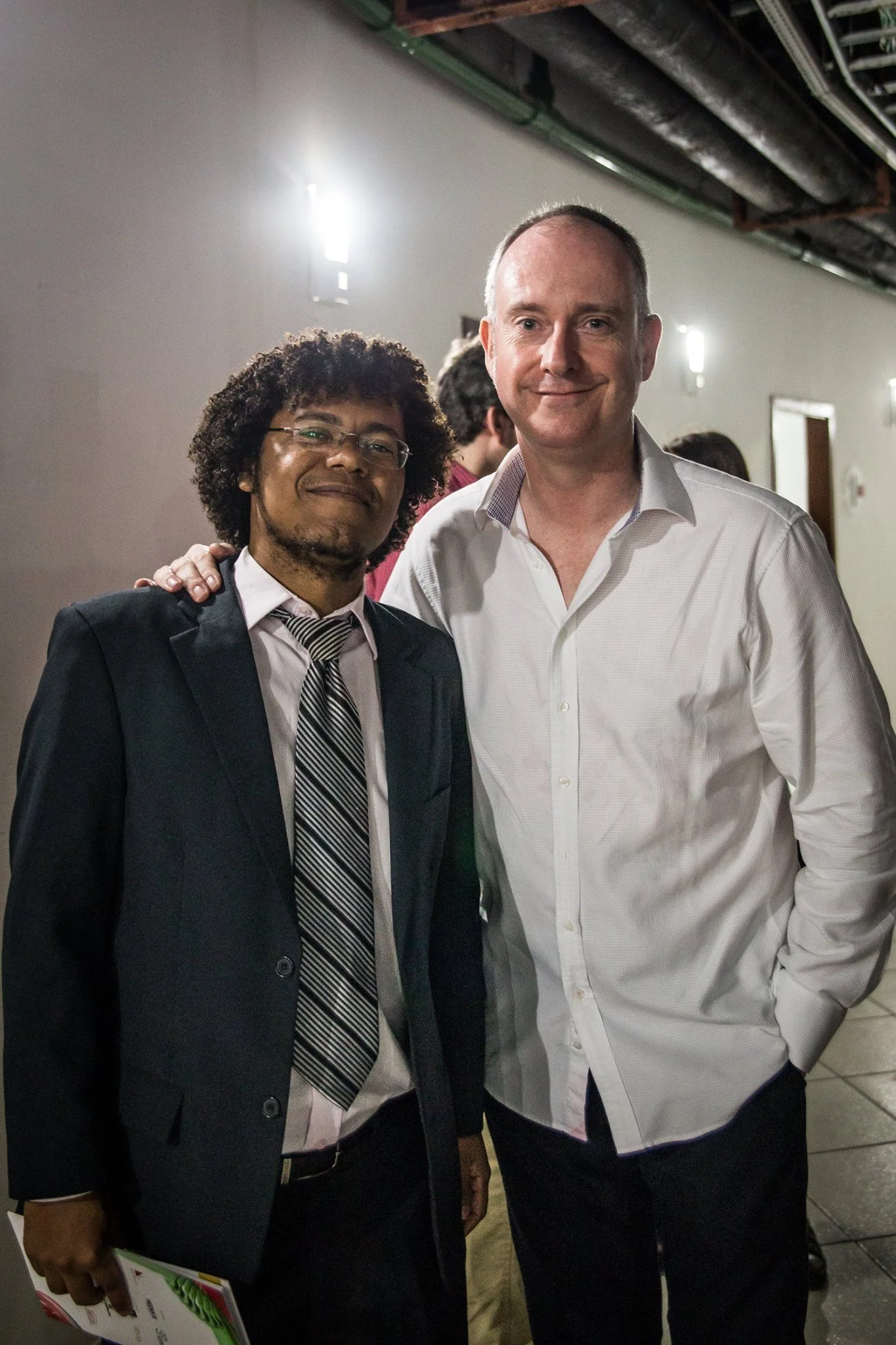 Two men standing together indoors, one wearing a black suit, striped tie, and glasses, and the other in a white dress shirt. The man in the suit is holding a brochure or magazine. They are smiling at the camera, with several other people in the background.
