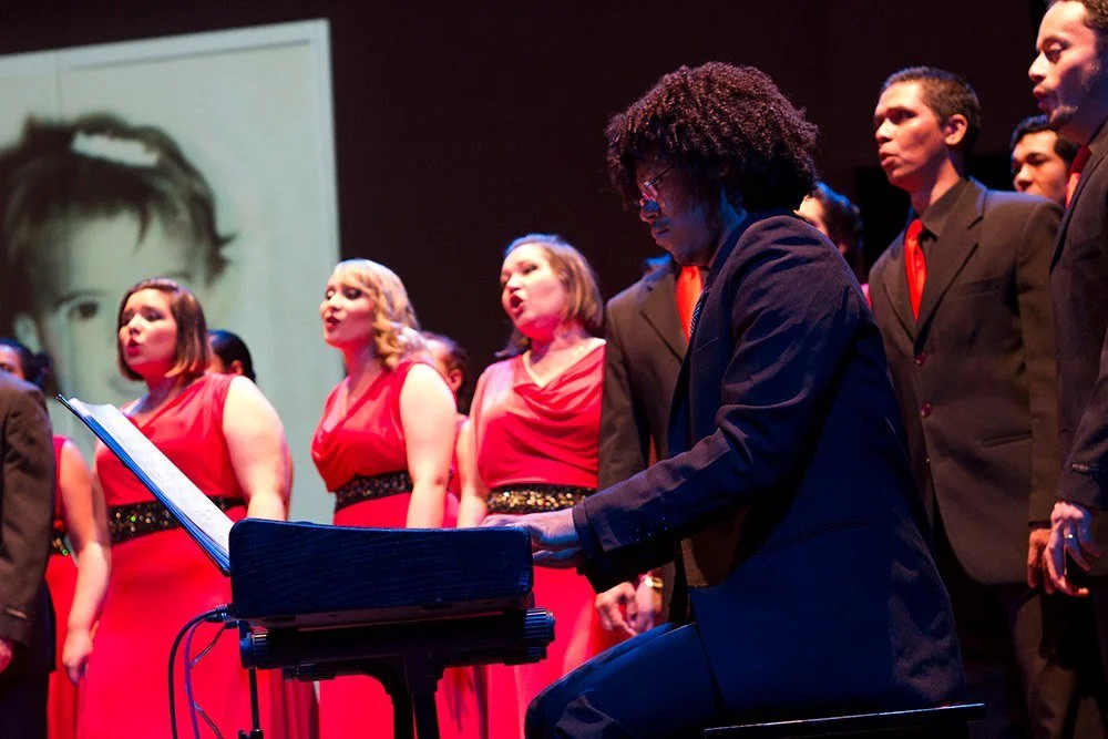 A choir performance with women in red dresses and men in suits, one person playing keyboard, on a stage with a large slideshow projection in the background.