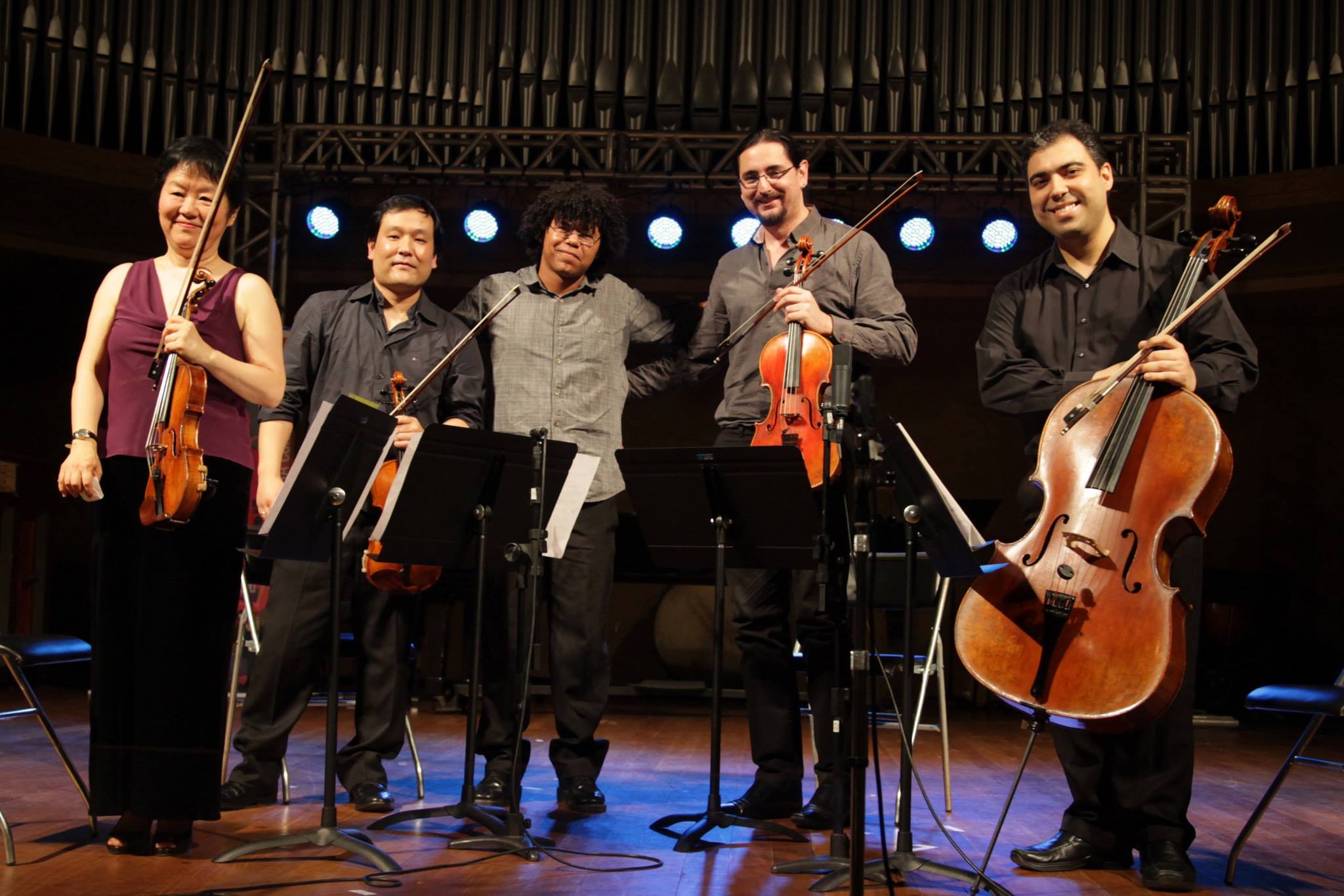 Group of five musicians on stage with classical instruments, standing in front of a stage with blue lights and metal framework, smiling after a performance.