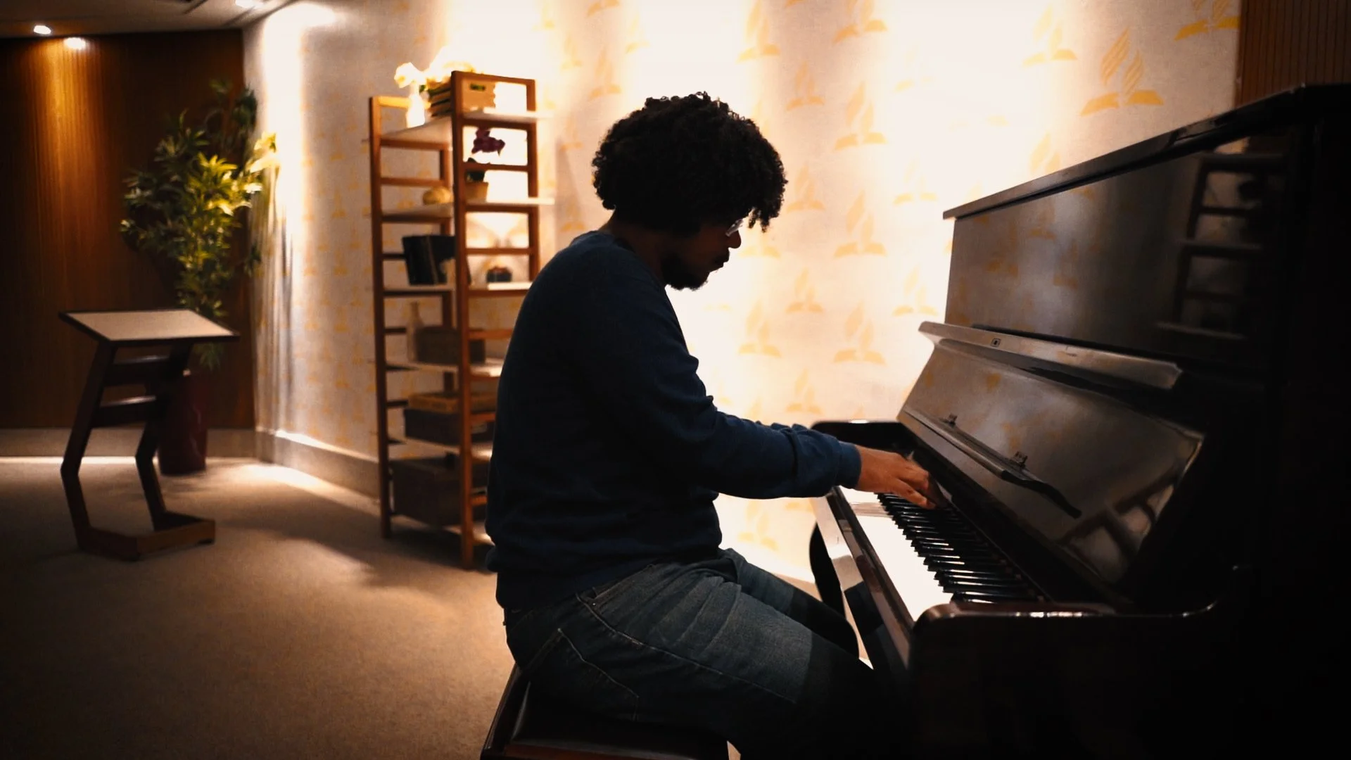 A person playing a black upright piano in a warmly lit room with patterned wallpaper, wooden shelves, and indoor plants.