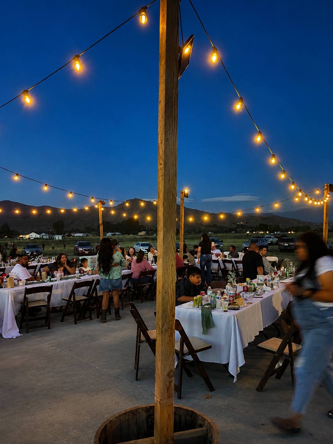 Outdoor evening celebration with long tables covered in white tablecloths, decorated with food and drinks, under string lights, with people socializing, mountains in the background, and a clear sky at dusk.
