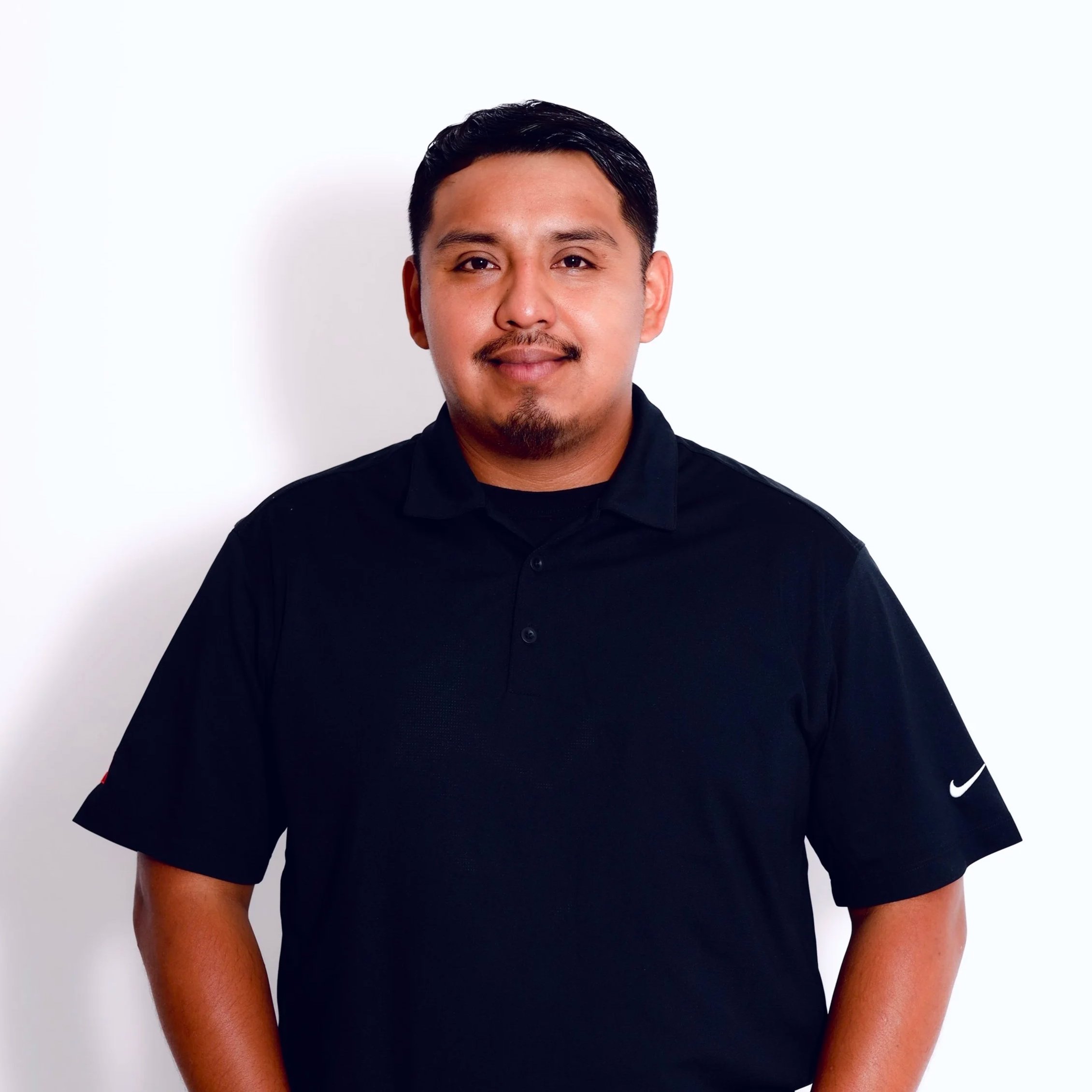 Close-up of a man with short black hair, a goatee, wearing a black Nike shirt, standing against a plain white background.