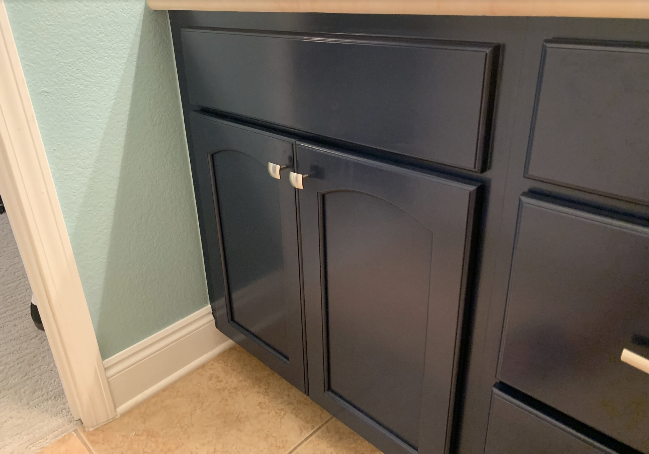 A black kitchen cabinet with two drawers and a door, positioned against a green wall with textured paint, next to a white door frame and beige tiled flooring.