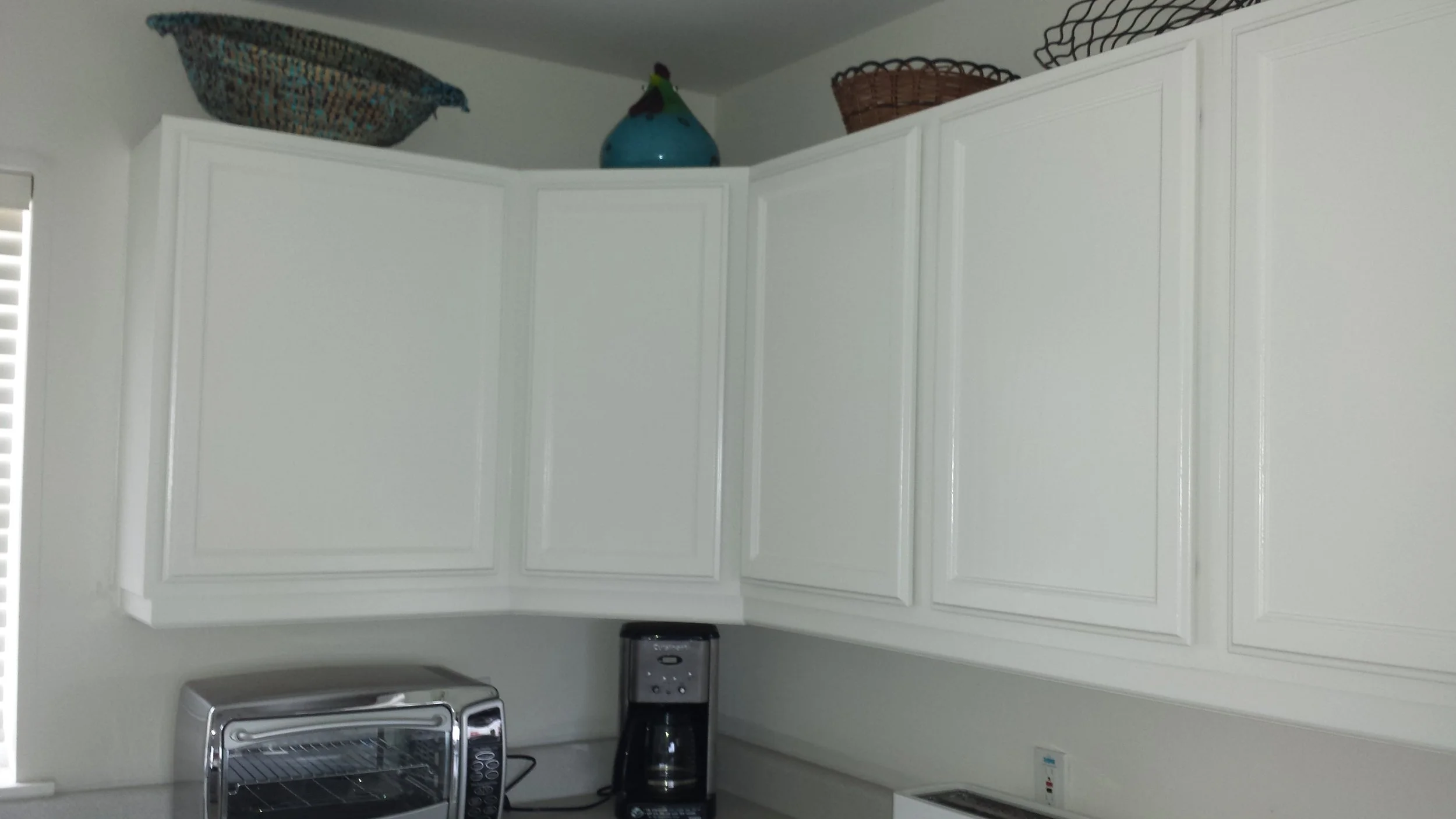 White kitchen cabinets with corner shelving holding baskets and vases, with small appliances and a coffee maker on the countertop.