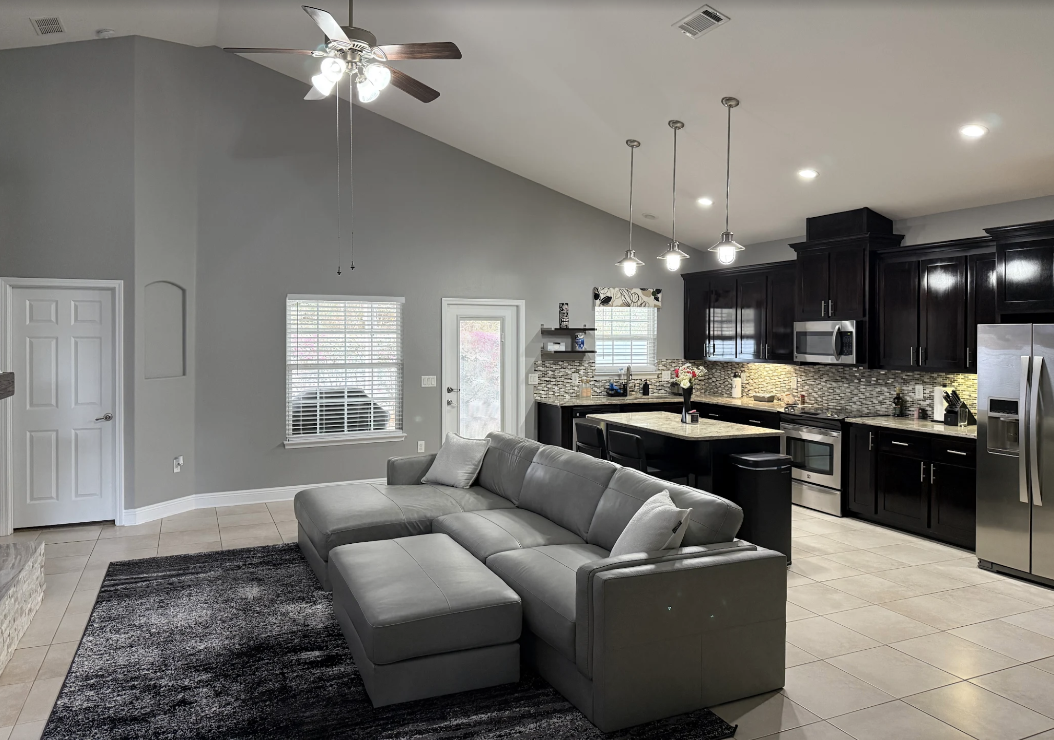 Bright open-concept kitchen and living room with gray walls, black cabinets, white countertops, stainless steel appliances, and a gray sofa. Kitchen features pendant lighting and a window with a floral valance.