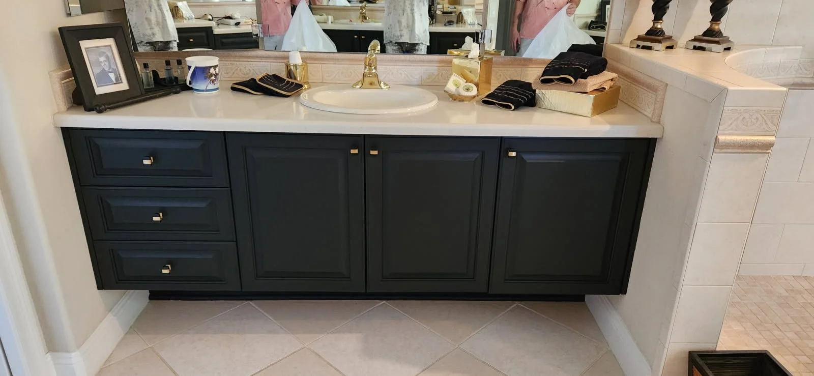 Bathroom vanity with black cabinets, white countertop, and gold fixtures, mirror above with reflection of the sink, and various toiletries and decorations.