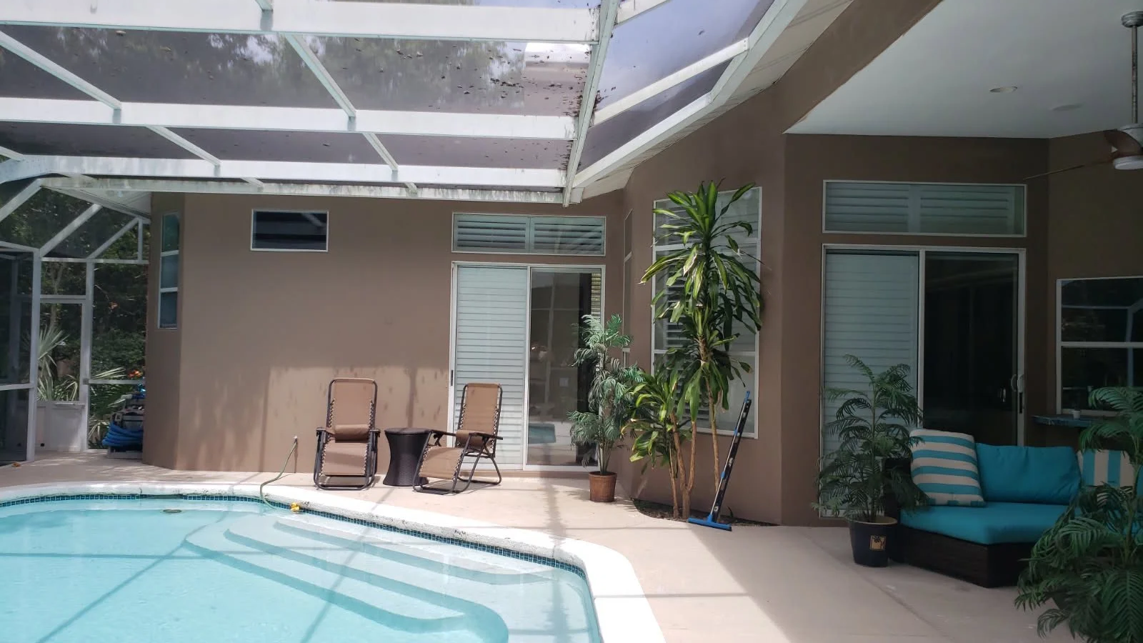 Outdoor patio with swimming pool, a glass enclosure, two chairs with a small table, and a blue couch with striped pillows, surrounded by potted plants and windows.