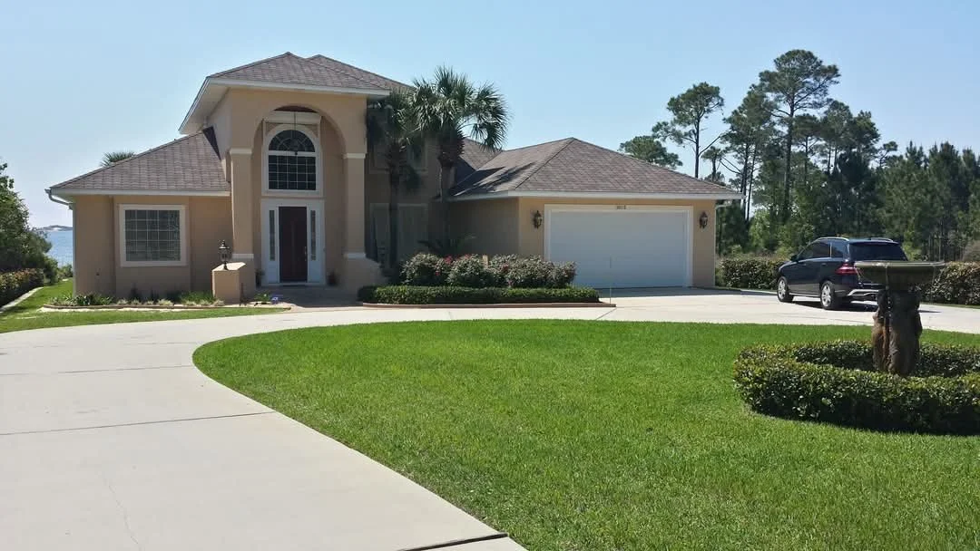 A large, modern house with a well-maintained lawn, driveway, and two parked cars in the driveway, with trees in the background.