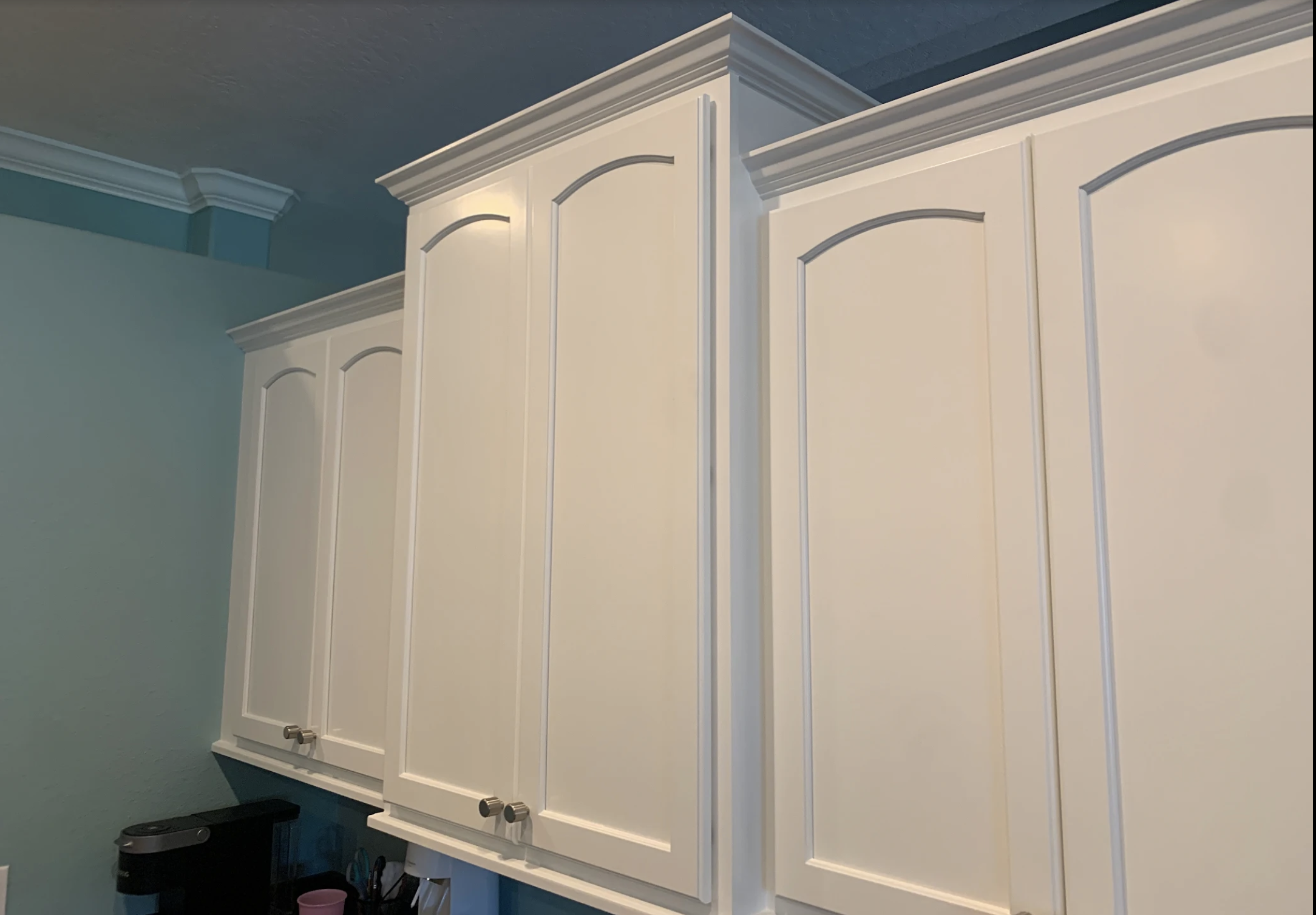 White kitchen cabinets with decorative molding, mounted on a teal wall.
