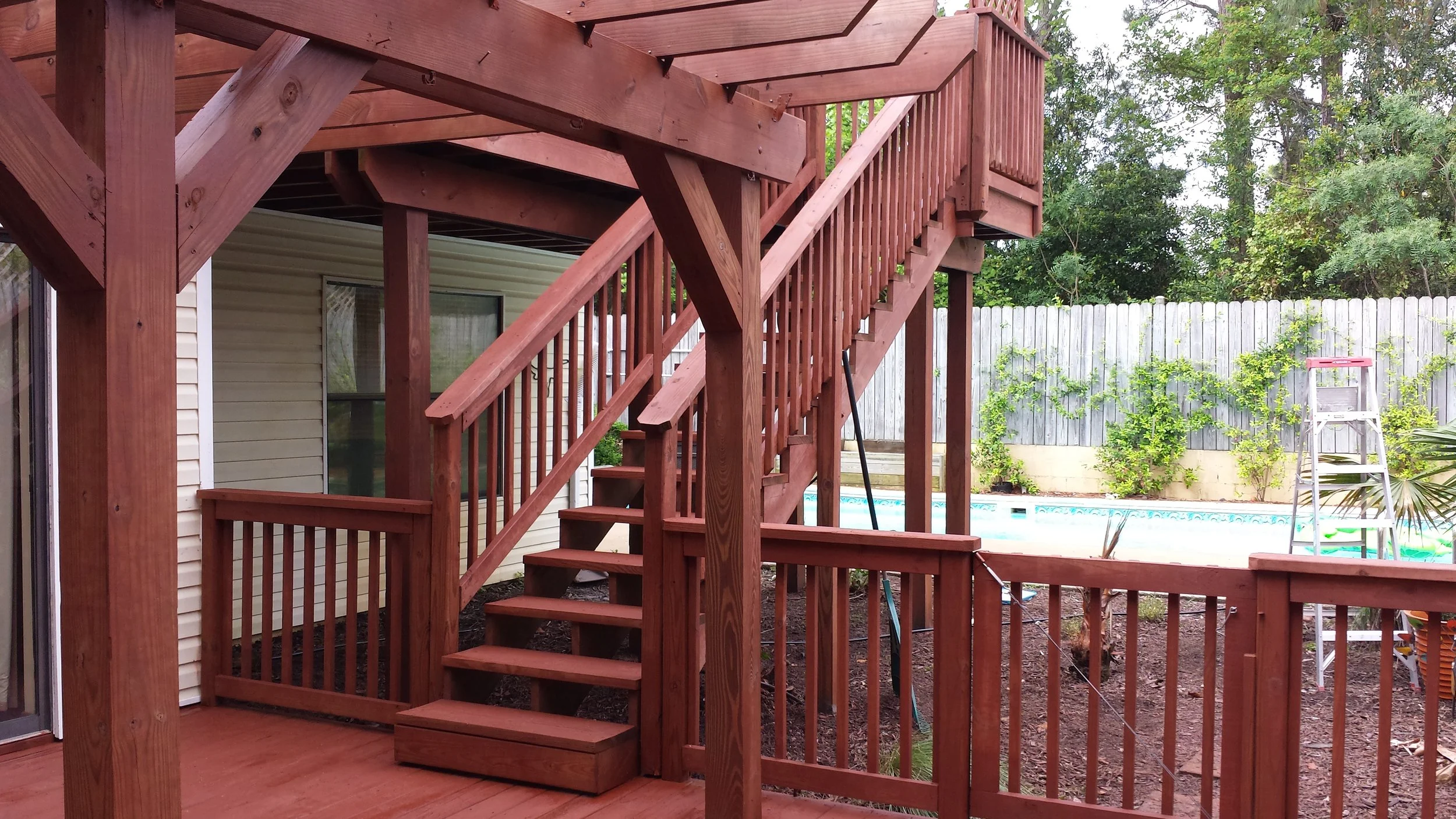 Backyard deck with stairs, wooden railing, and a pool in the background, surrounded by trees and a wooden fence.