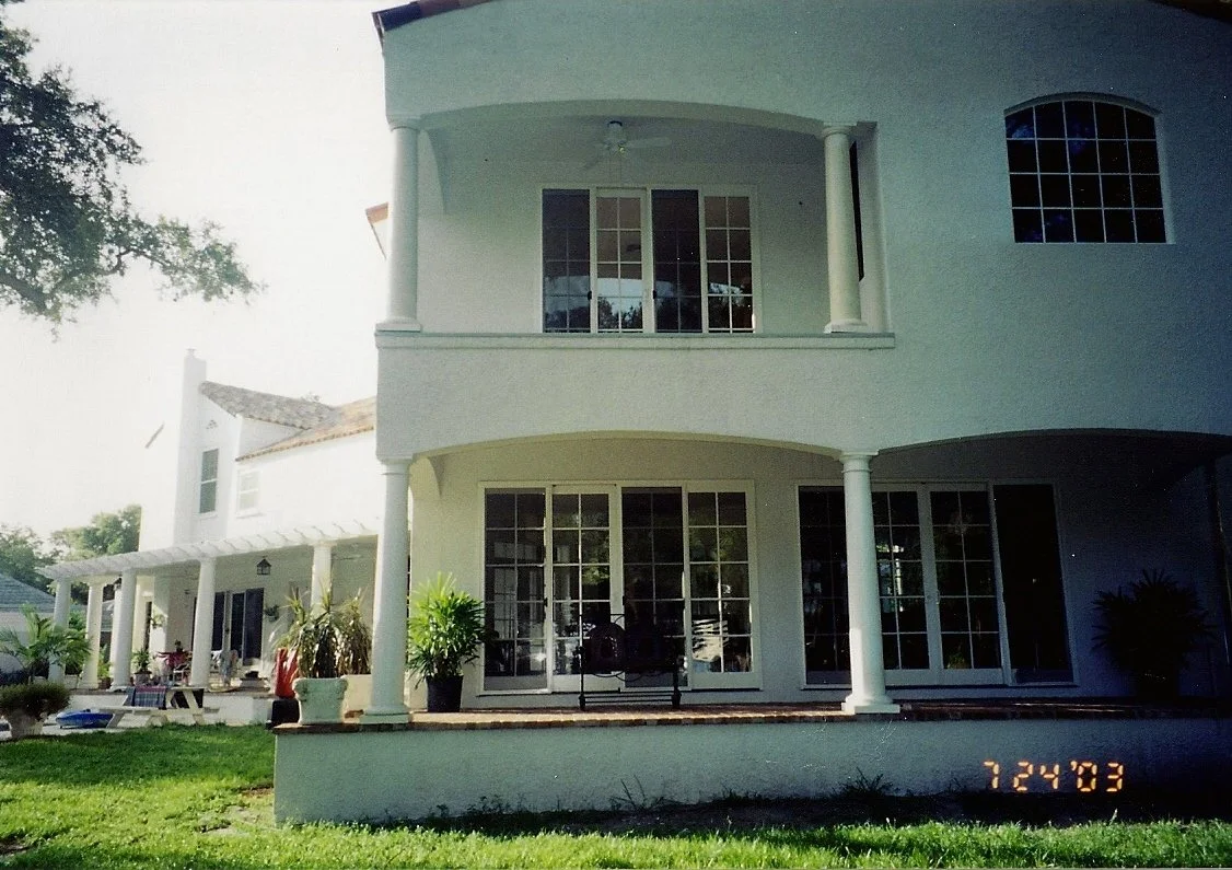 Rear view of a two-story white house with large windows and porch, surrounded by green grass and plants, on a sunny day.