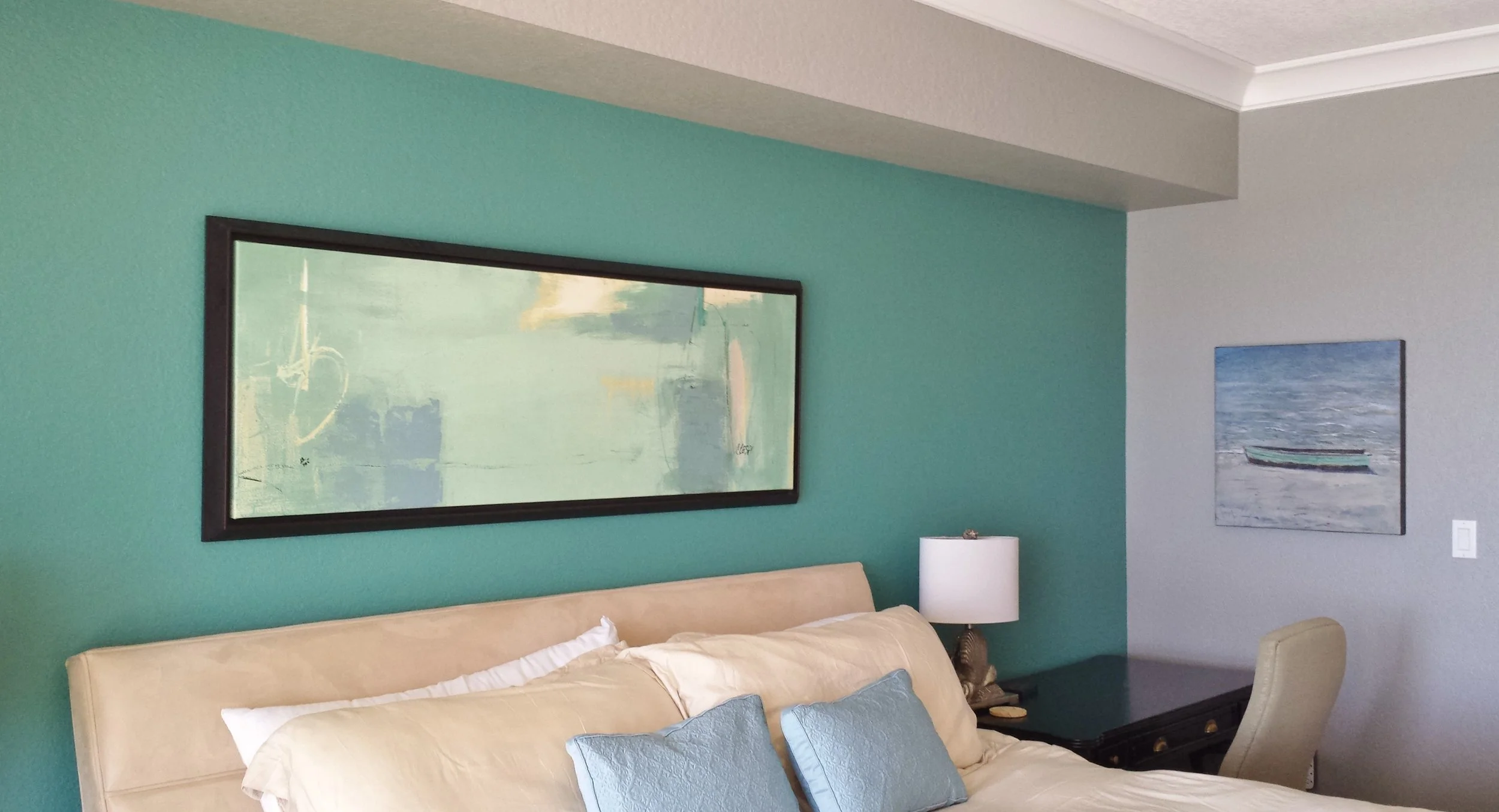 A bedroom with a turquoise accent wall with two framed art pieces, one above the bed and one on the adjacent wall, a beige upholstered headboard, pillows, a white bedside lamp, and a dark dresser.