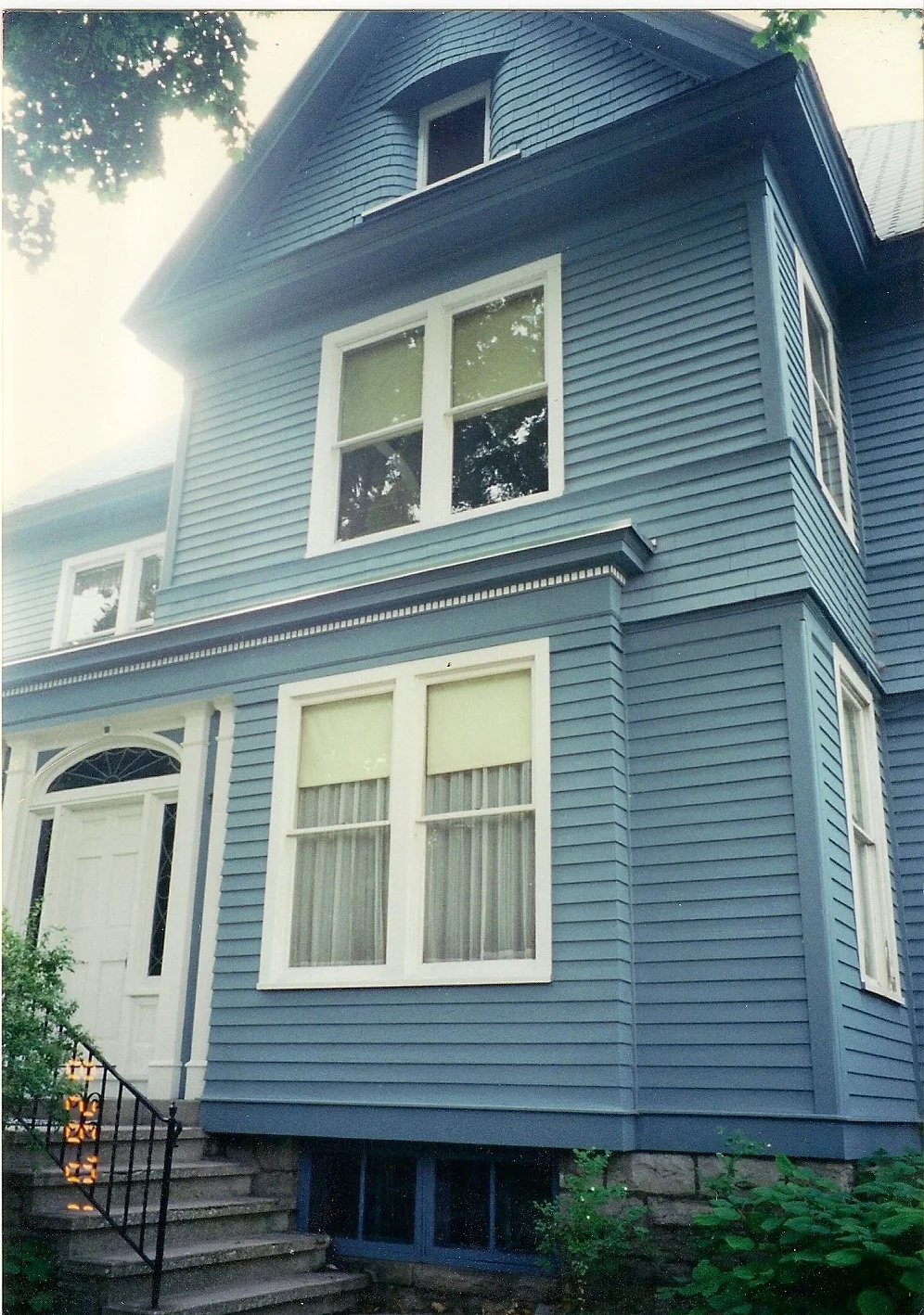 A blue, multi-story house with white window frames, front steps, and black railings, surrounded by greenery.