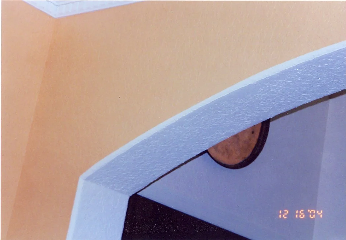 Close-up of a ceiling corner with a gold-colored circular wall decor. The ceiling has a textured White paint, and the wall is painted in a warm yellow-orange color.