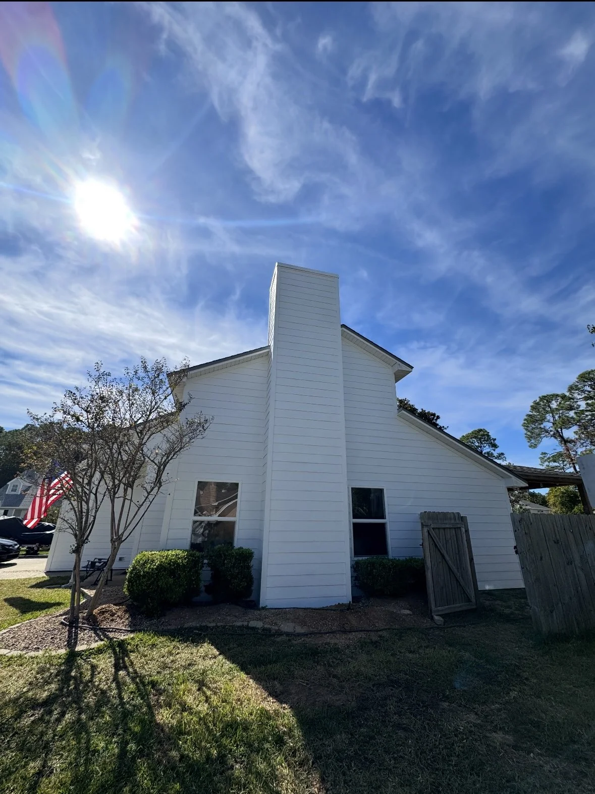 White house with a chimney, trees, a small garden area, and an American flag in front on a sunny day with a blue sky and some clouds.