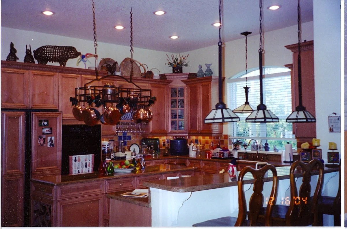 A kitchen with wooden cabinets, hanging copper pots, a pot rack, and decorative items on top of cabinets. There are three pendant lights over the counter, a window with blinds, and various kitchen appliances and decorations on the countertops.