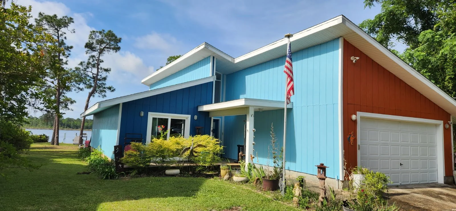Colorful house with blue and red siding, white garage door, American flag, surrounded by green lawn and trees near a body of water.