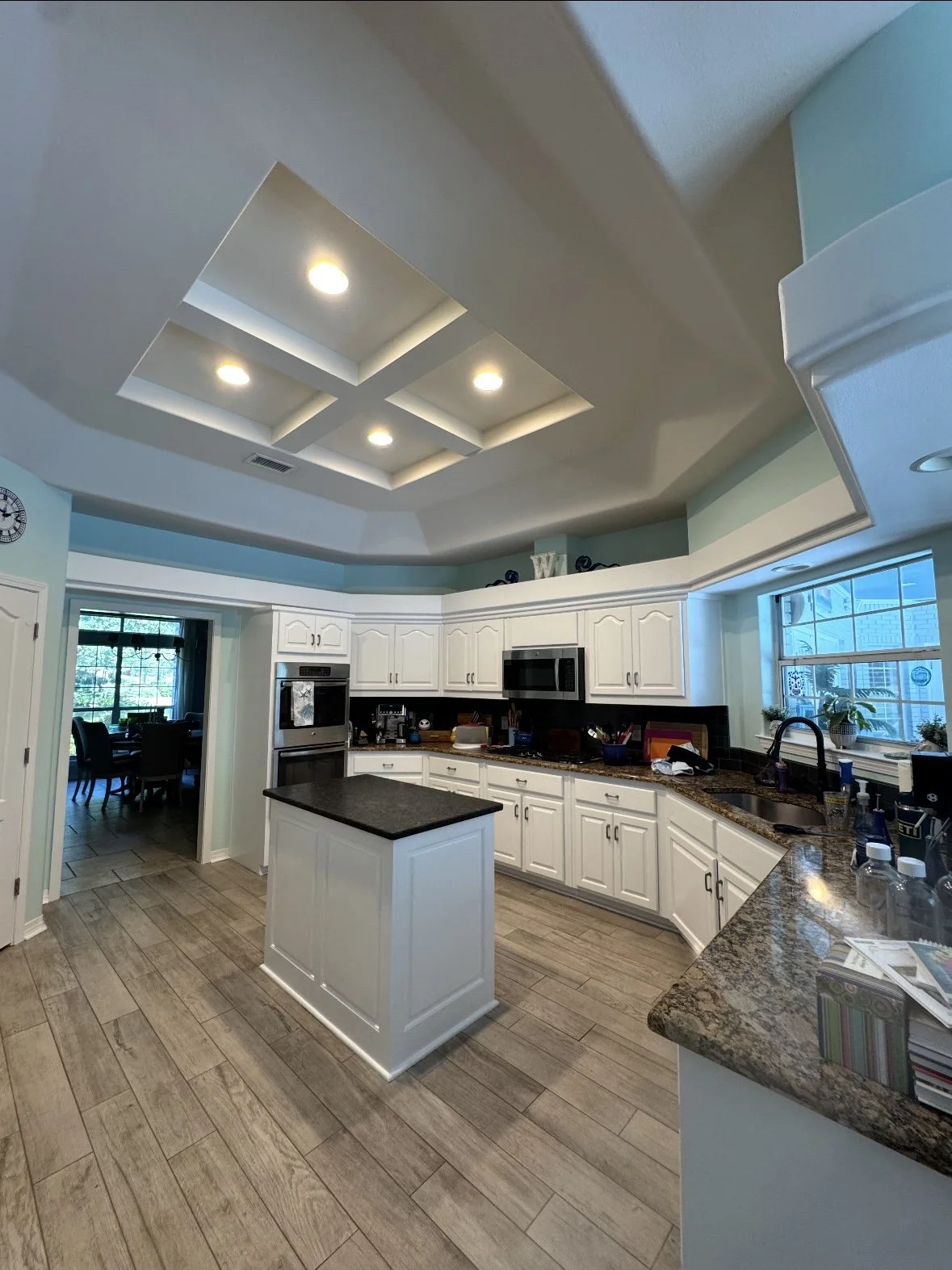 Bright kitchen with white cabinets, granite countertops, and a small island. Recessed ceiling lights and large window above the sink. Visible doorway leading to dining area.