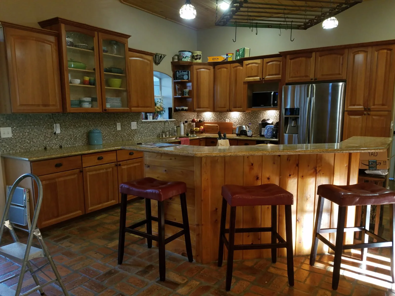 A kitchen with wooden cabinets, a granite countertop, a mosaic tile backsplash, stainless steel refrigerator, microwave, and countertop appliances. Three bar stools with leather seats are at the breakfast bar, and a step ladder is on the left side. T