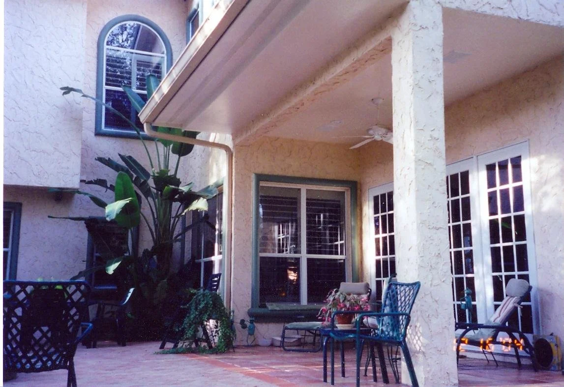 Back patio of a house with outdoor chairs, a large potted plant, and a window with white blinds, with a stucco wall and beige pillars.