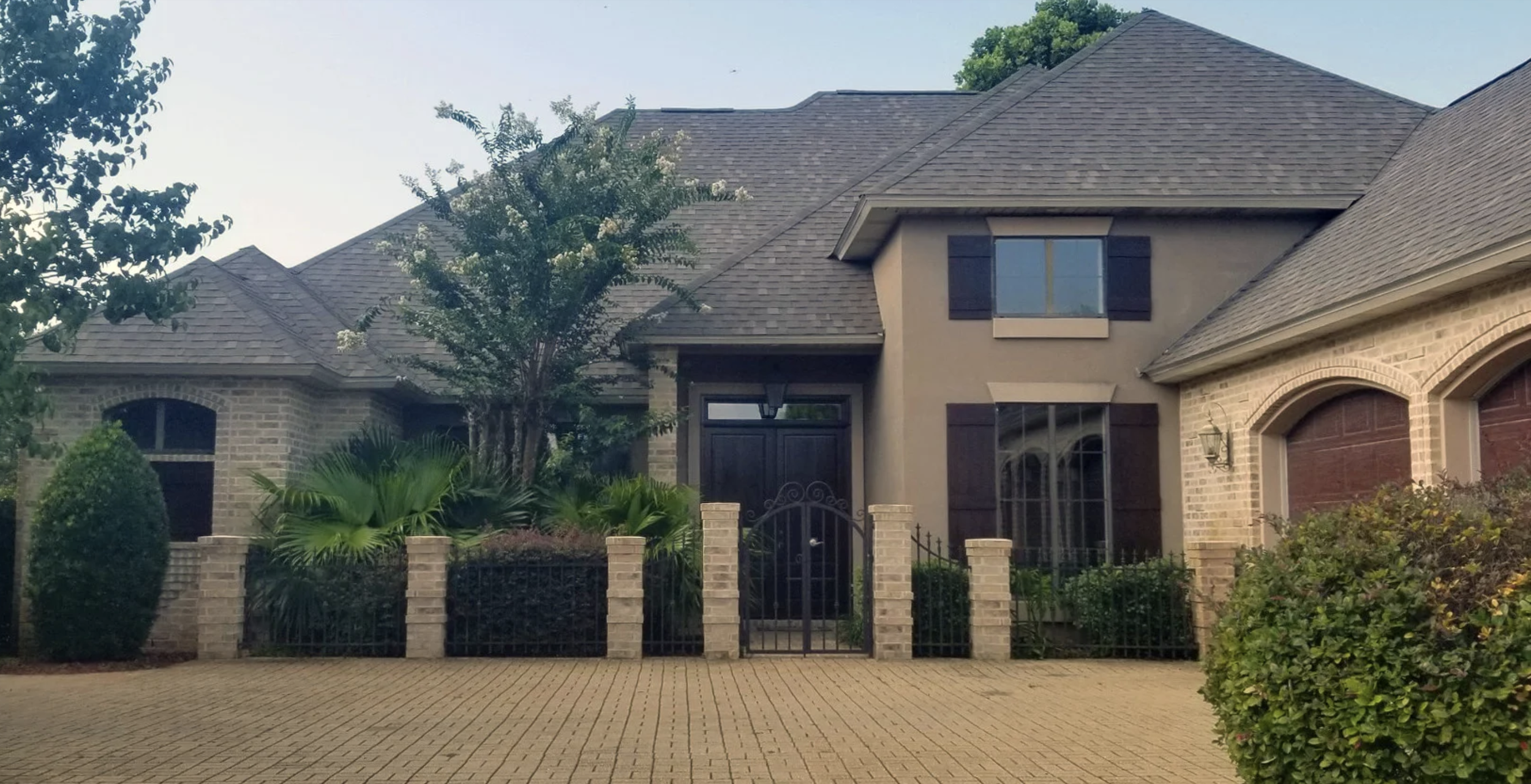 A large residential house with a brick and stucco exterior, multiple gabled roofs, a front yard with plants and trees, a black iron gate, and a driveway.
