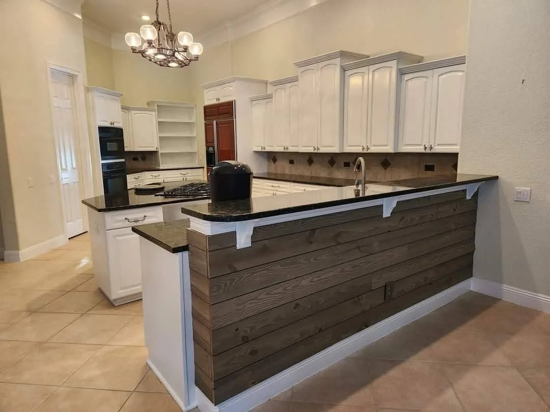 A kitchen with white cabinetry, a dark granite countertop, and a two-tone wooden accent on the kitchen island. There is built-in oven and microwave, open shelves, a faucet, and a chandelier overhead. The floor is tiled in a beige color.
