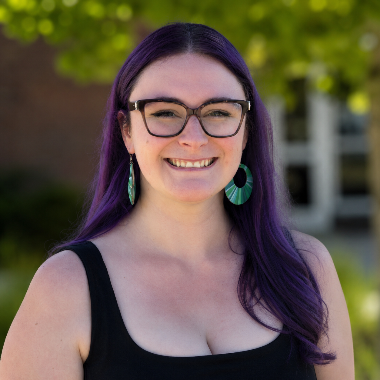 A picture of Madeline Leigh Jones, who is a woman with long purple hair, wearing glasses and green earrings, smiling outdoors with trees and a building in the background.