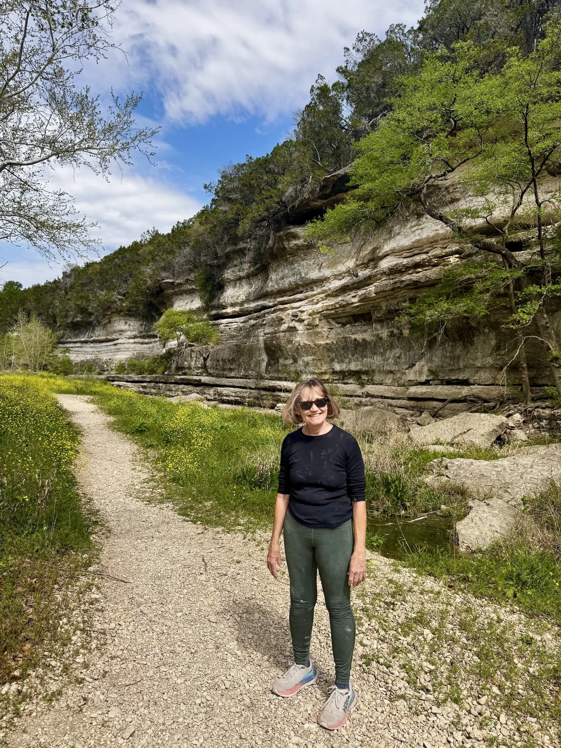 A smiling woman in sunglasses and athletic clothing standing on a gravel trail in a natural park setting, with layered rock formations, trees, green foliage, and a partly cloudy blue sky in the background.