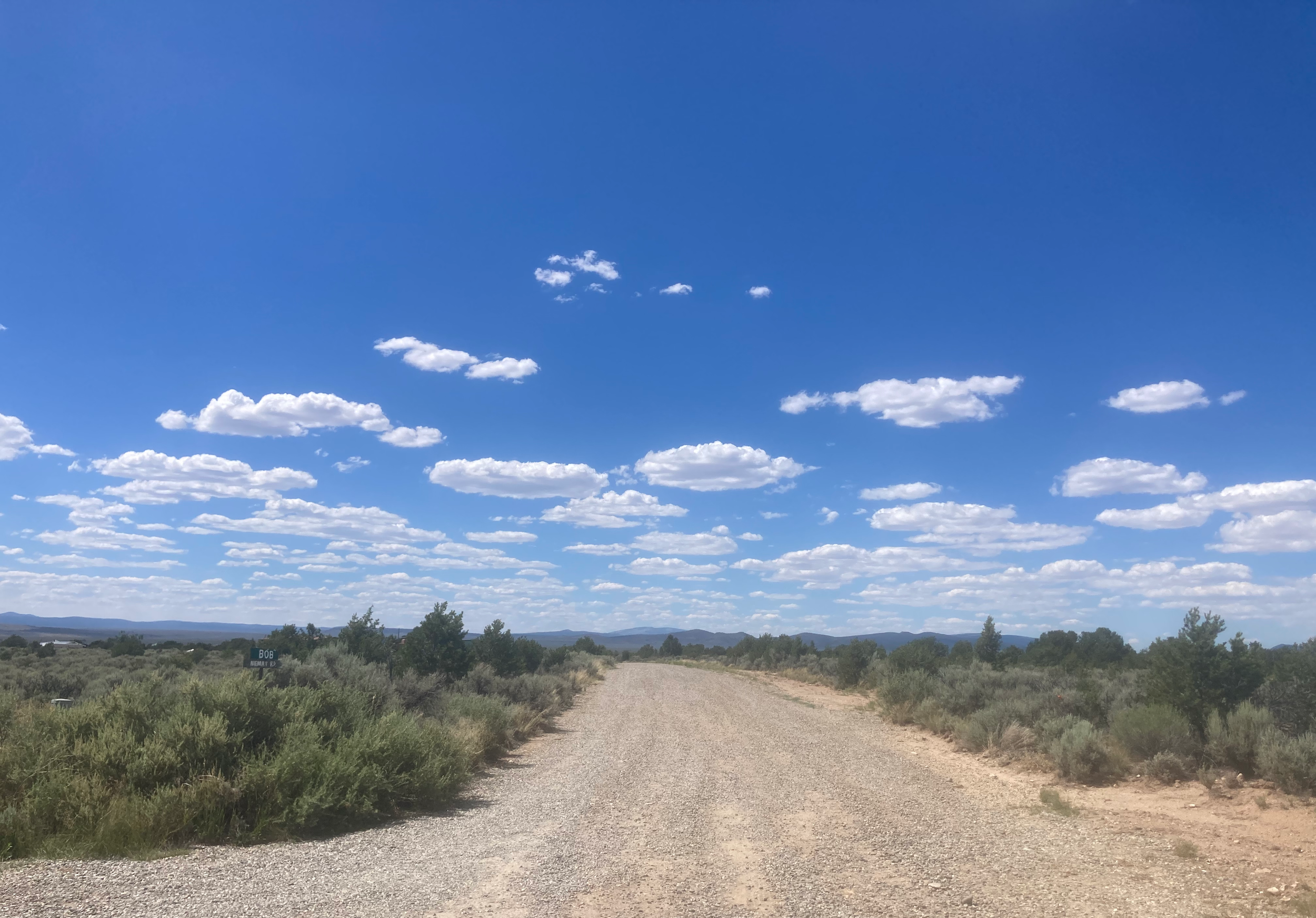 A dirt road in a rural area with green bushes and trees on either side, leading towards mountains in the distance under a blue sky with scattered white clouds.